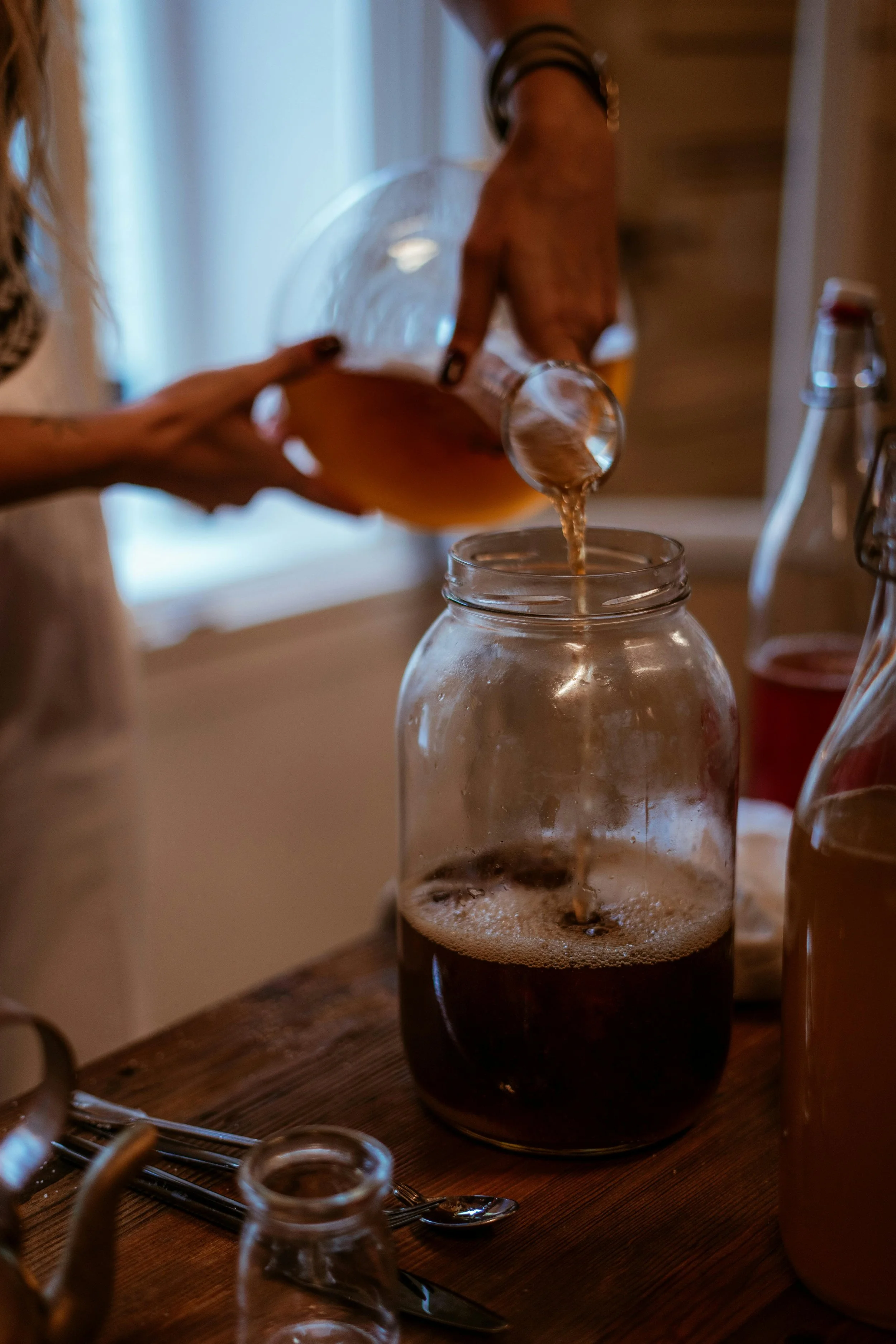 Person pouring a dark beverage into a glass jar, with other jars and utensils on a wooden table.