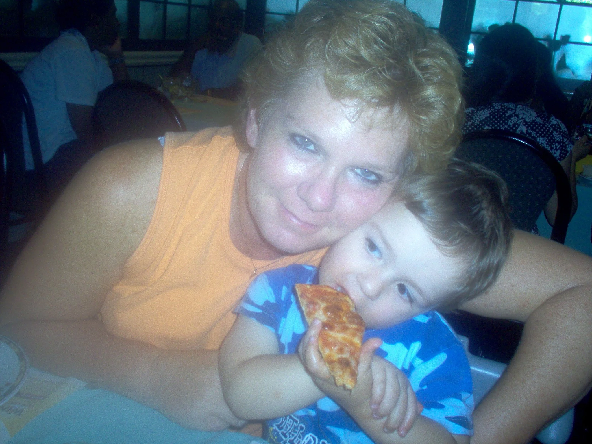 A woman with short blonde hair and blue eyes hugging and showing affection towards a young boy with light skin and short brown hair, who is eating a slice of pizza. They are sitting at a table in a restaurant with other people in the background.