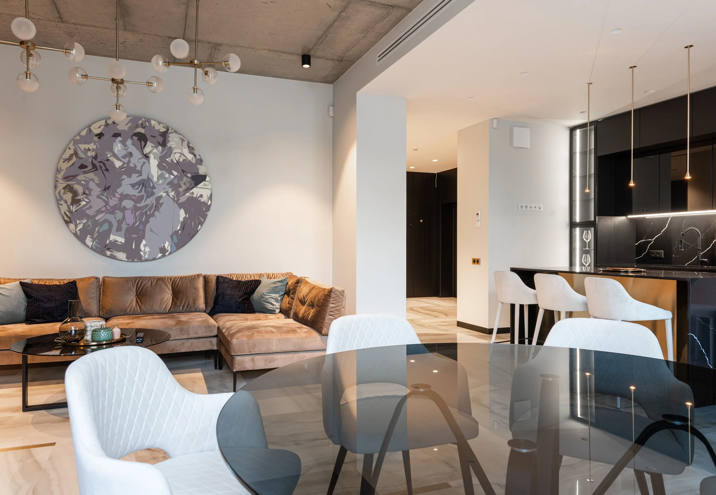 Modern living room and kitchen with a beige sofa, abstract wall art, white chairs around a black glass dining table, and a kitchen area with black cabinetry, marble countertops, and pendant lighting.