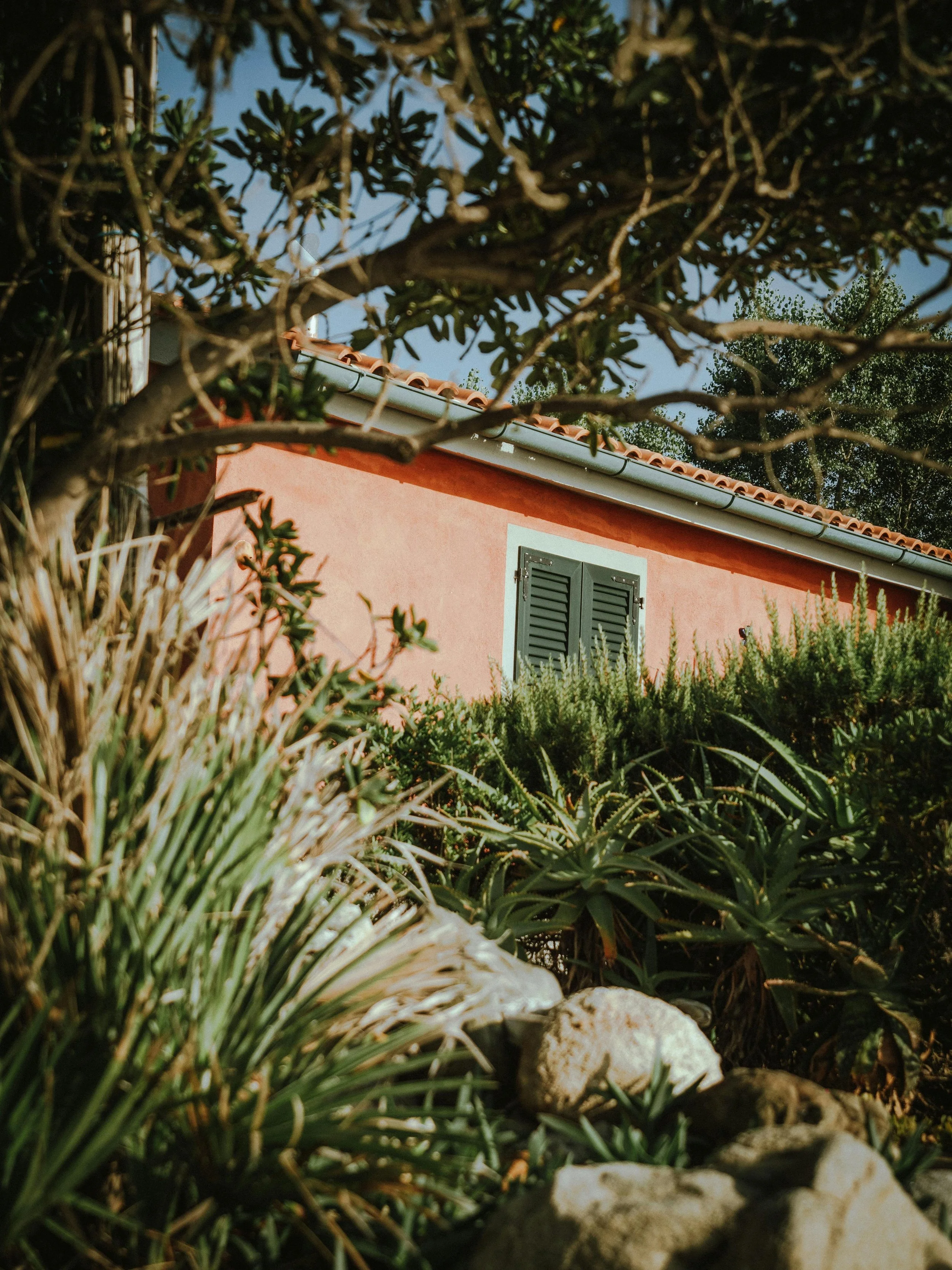 A pink house with a gray roof and closed green shutters, viewed through lush greenery and rocks in the foreground.