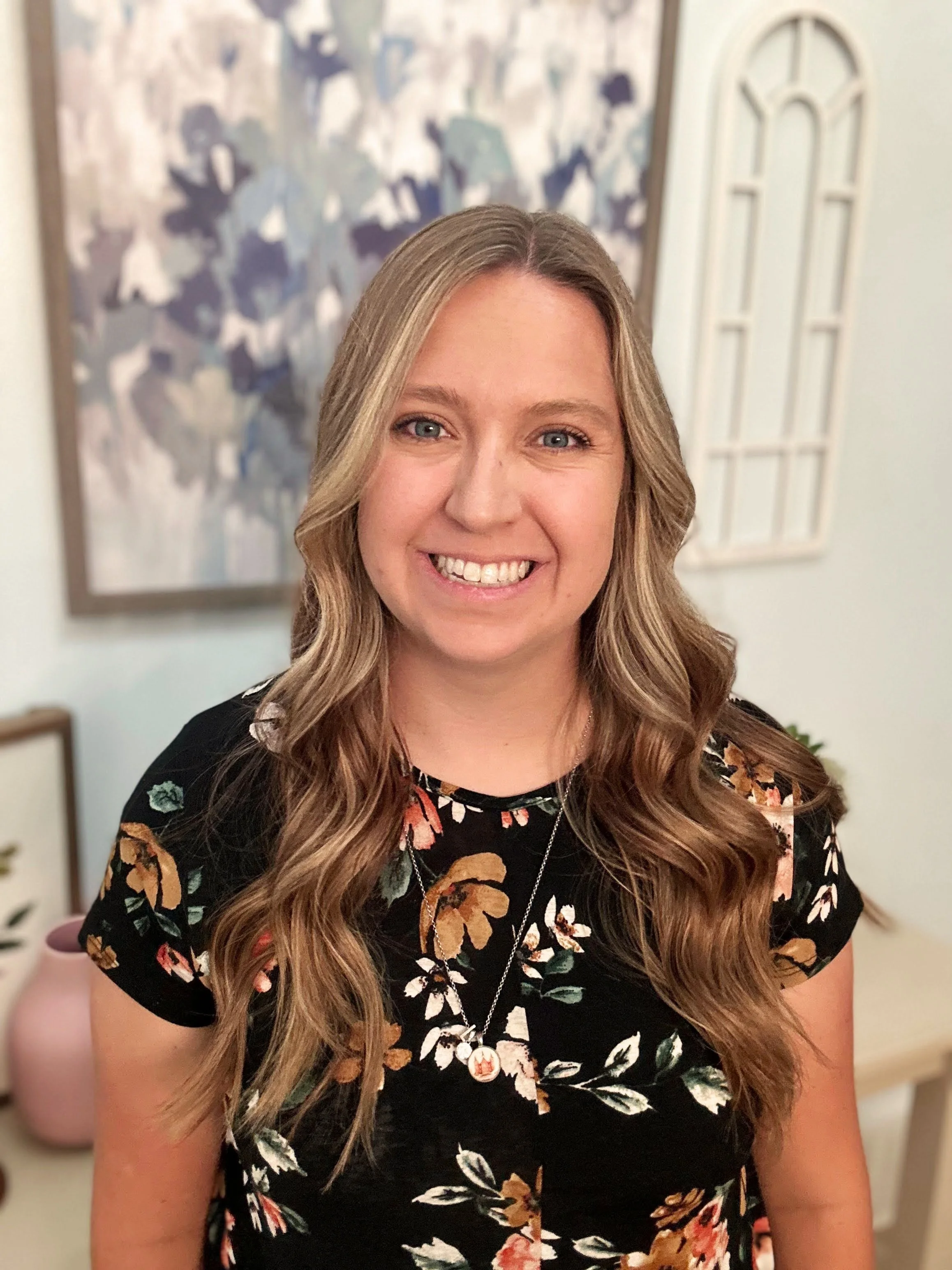 A woman smiling, with long wavy brown hair, wearing a floral dress and a necklace, standing indoors with wall art and a pink vase behind her.