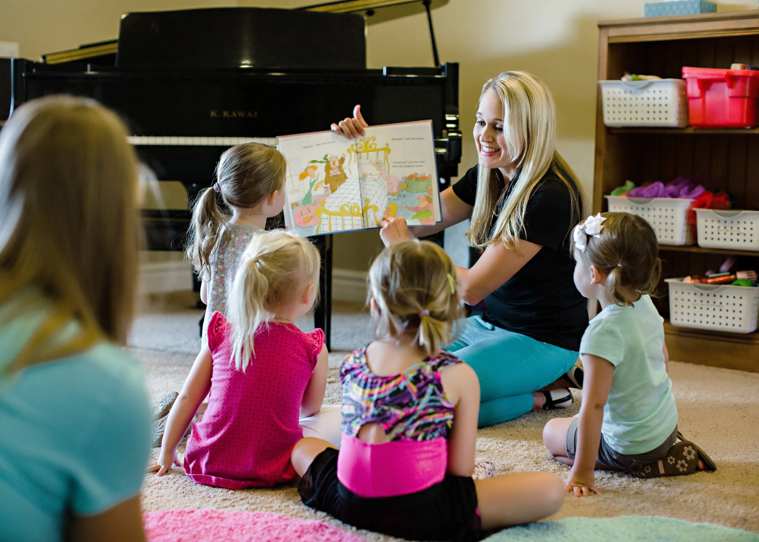 A woman reading a colorful picture book to a group of six young girls sitting on the carpet in a room with a black grand piano and shelves filled with baskets of toys.