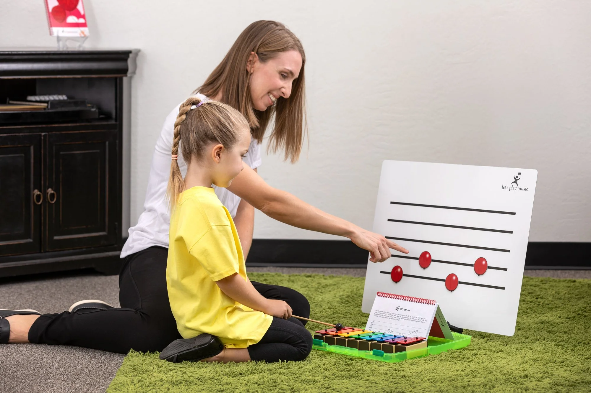 A woman and a girl sitting on a green rug, playing music with a xylophone and a music staff board, smiling and looking at the music stand.