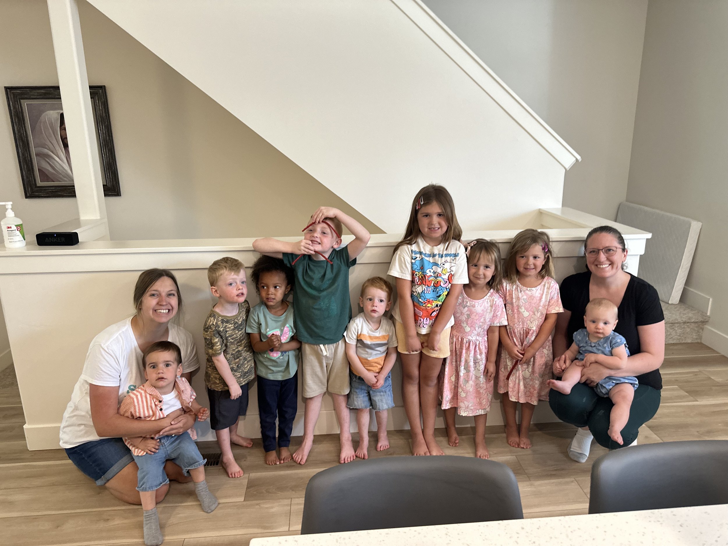 Group of ten children and two women posing inside a house, with a staircase in the background, smiling and standing near a half-wall.