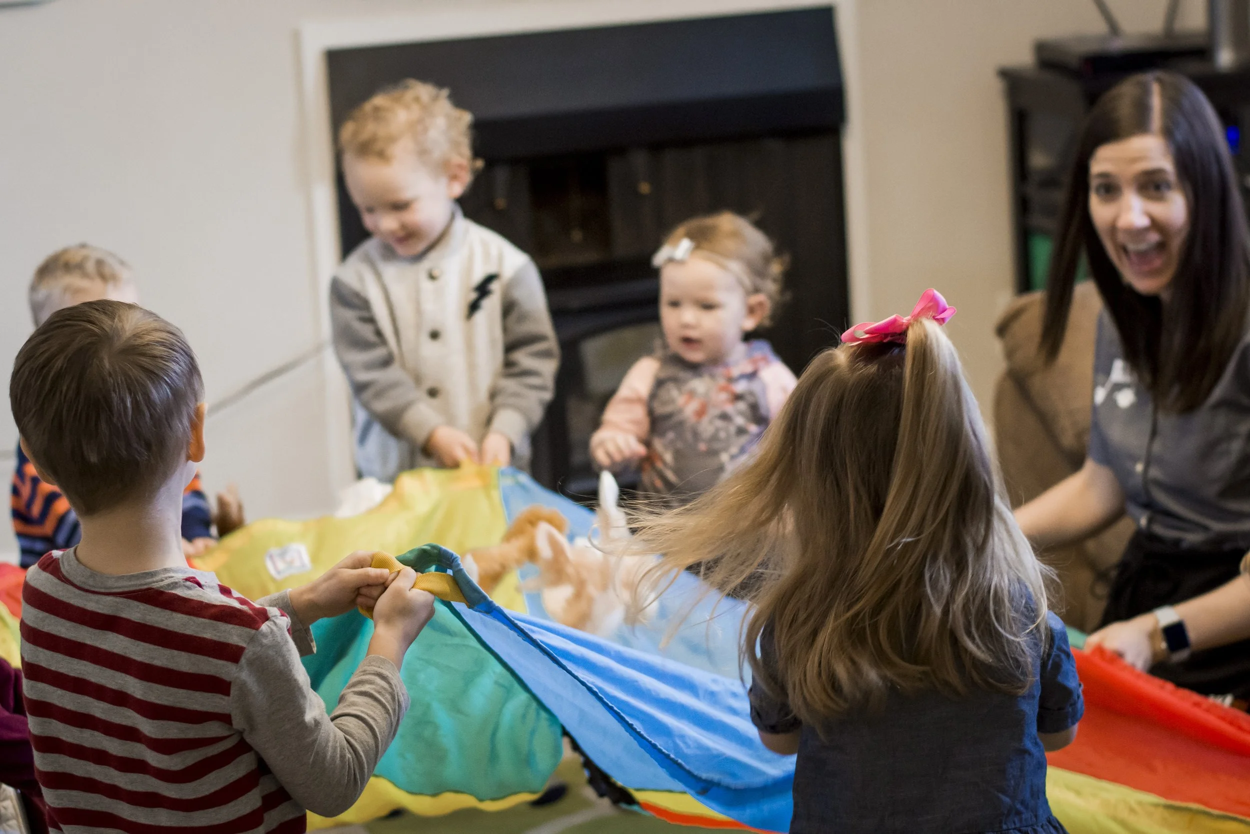 Children and an adult engaging in a parachute game indoors.