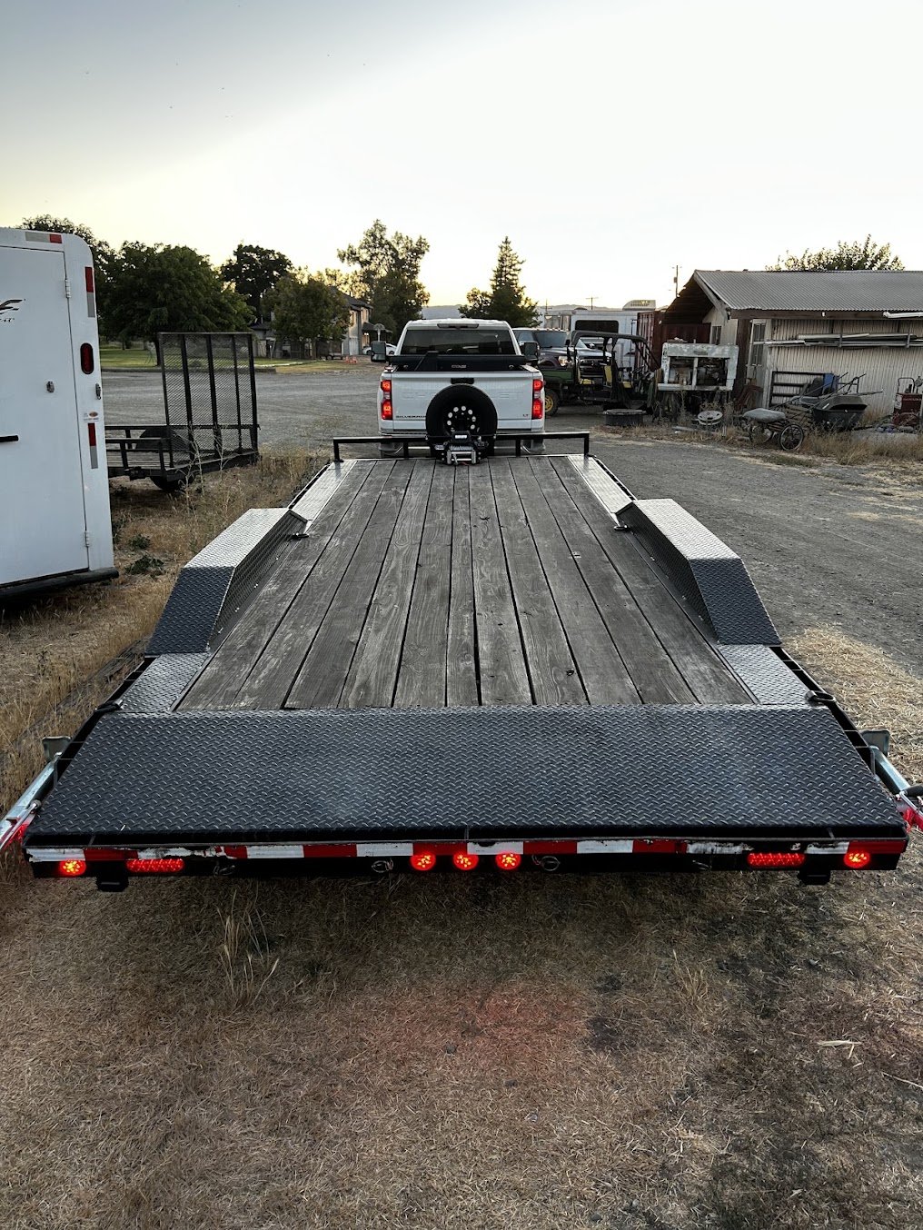 A flatbed trailer with wooden planks, parked outdoors near a white truck and other equipment, with trees and a building in the background at sunset.