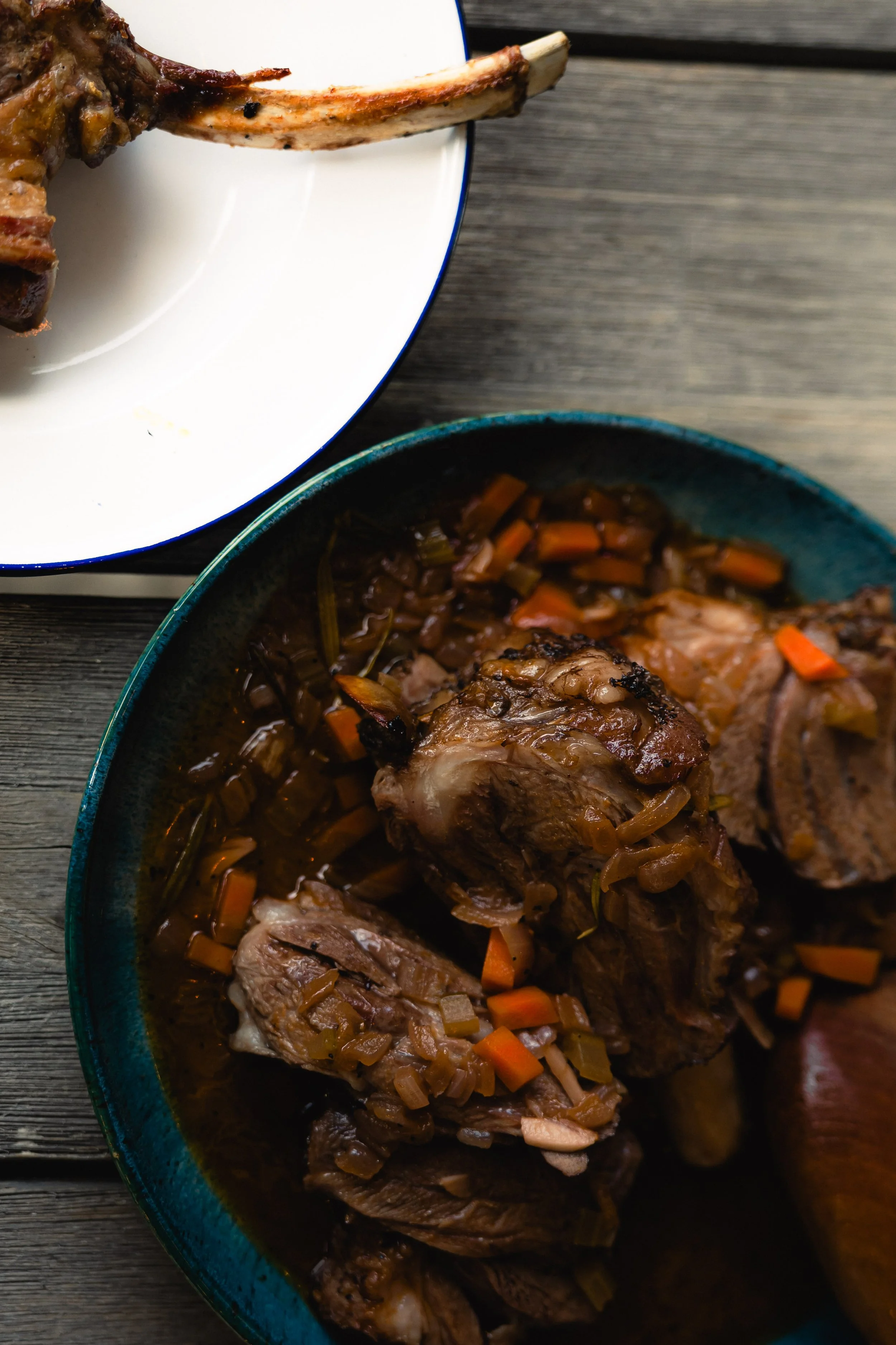 Braised lamb shanks with vegetable stew in a rustic bowl on a wooden surface, and a plate with a lamb chop for a farm to table lifestyle.