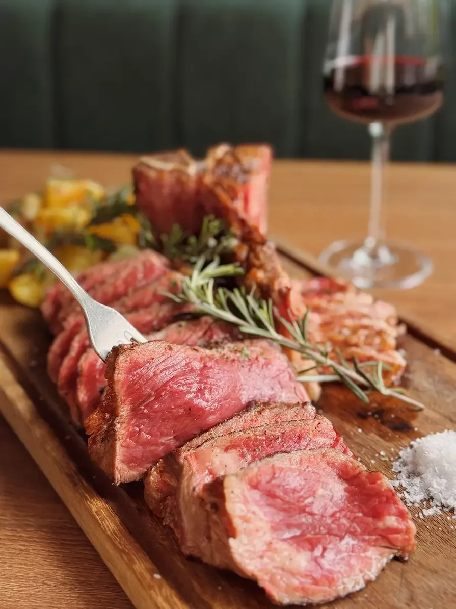 Sliced cooked steak on a wooden serving board with a sprig of rosemary, salt, and a glass of red wine in the background.
