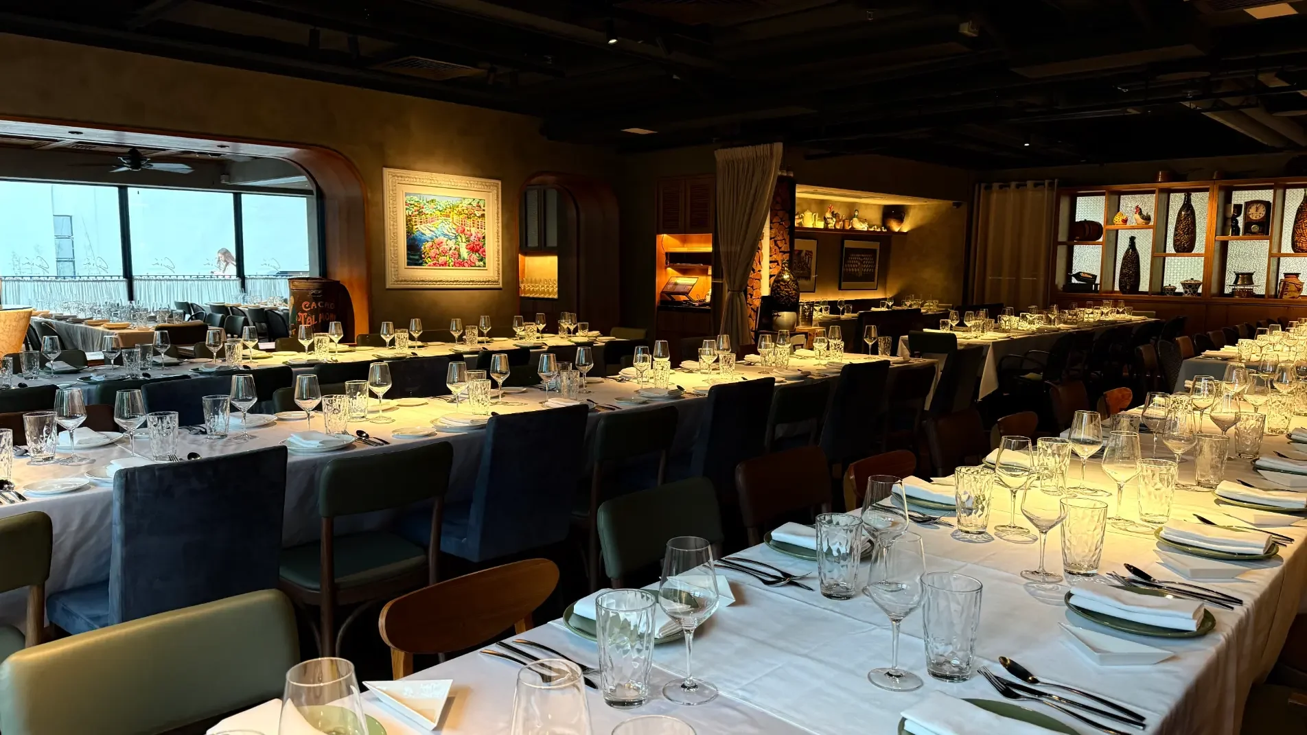 Elegant restaurant dining area with long tables set with white tablecloths, glassware, plates, and silverware, featuring a cozy and warm ambiance with artwork and decorative shelves.