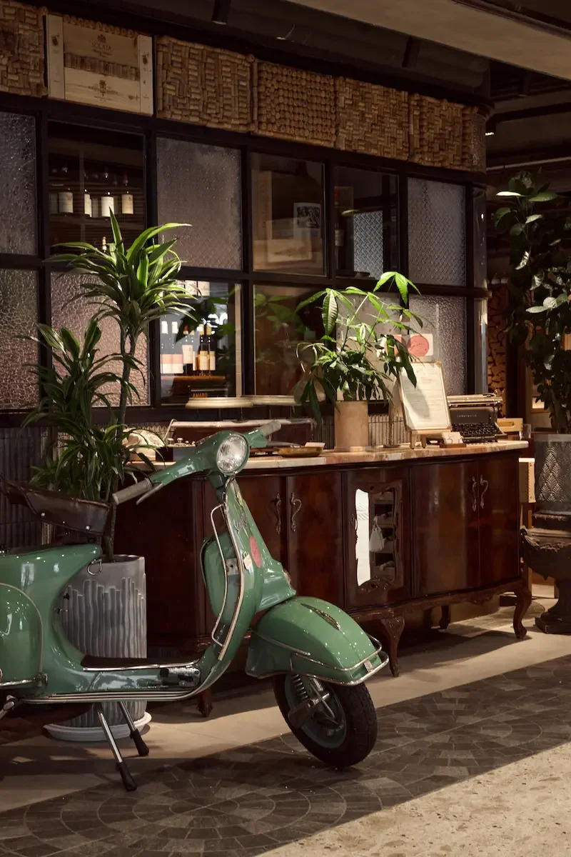 Vintage green scooter parked indoors near a wooden cabinet with potted plants on top, background featuring a glass partition, furniture, and decorative objects.