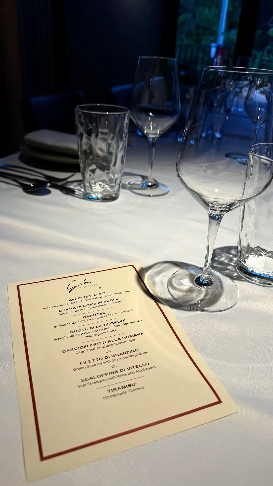 A restaurant table set with glassware, cutlery, and a menu on a white tablecloth, with a dimly lit background showing some outdoor scenery.