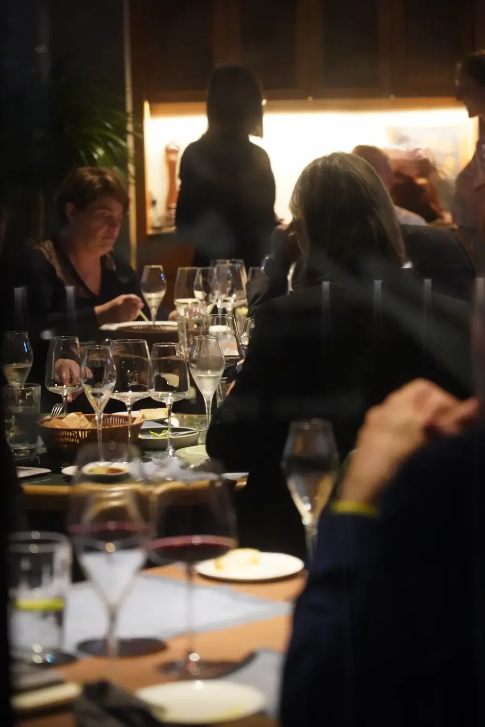 Group of people dining at a restaurant with wine glasses and plates on the table, in a dimly lit setting.