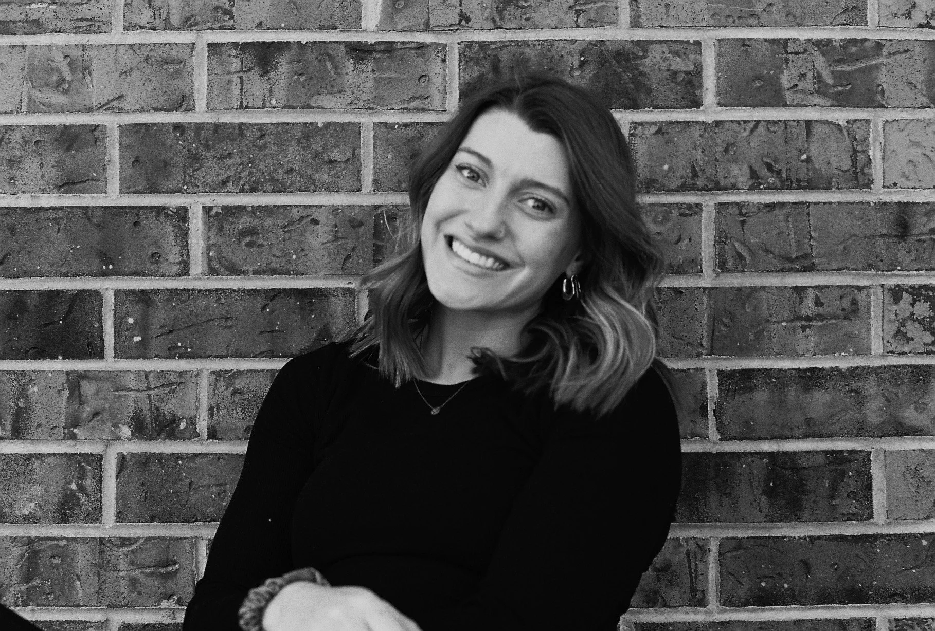 A young woman with shoulder-length wavy hair, smiling, in front of a brick wall. She is wearing earrings, a necklace, and a black long-sleeve shirt.