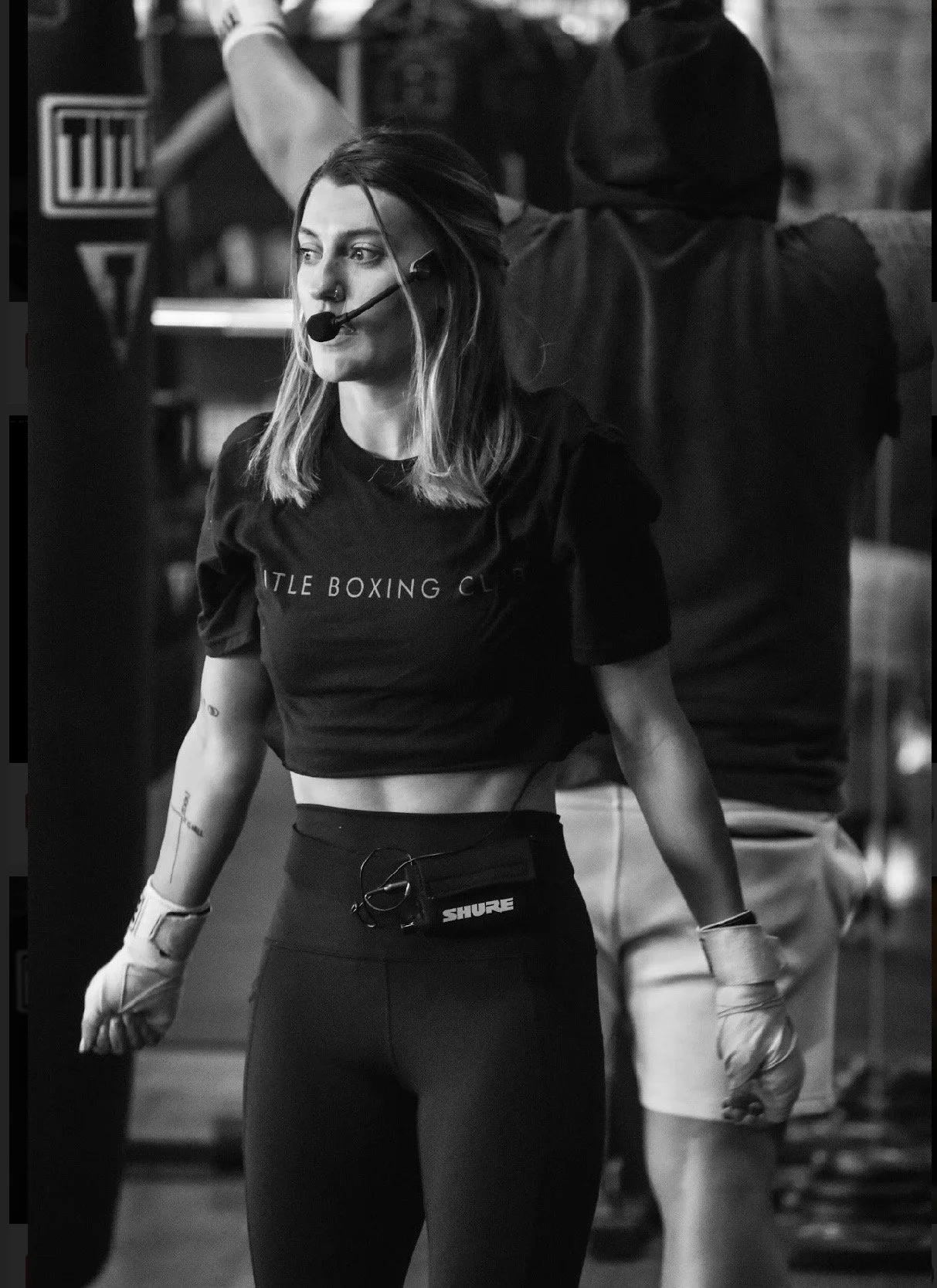 A woman with a headset and gloves, wearing a t-shirt and leggings, stands in a gym.