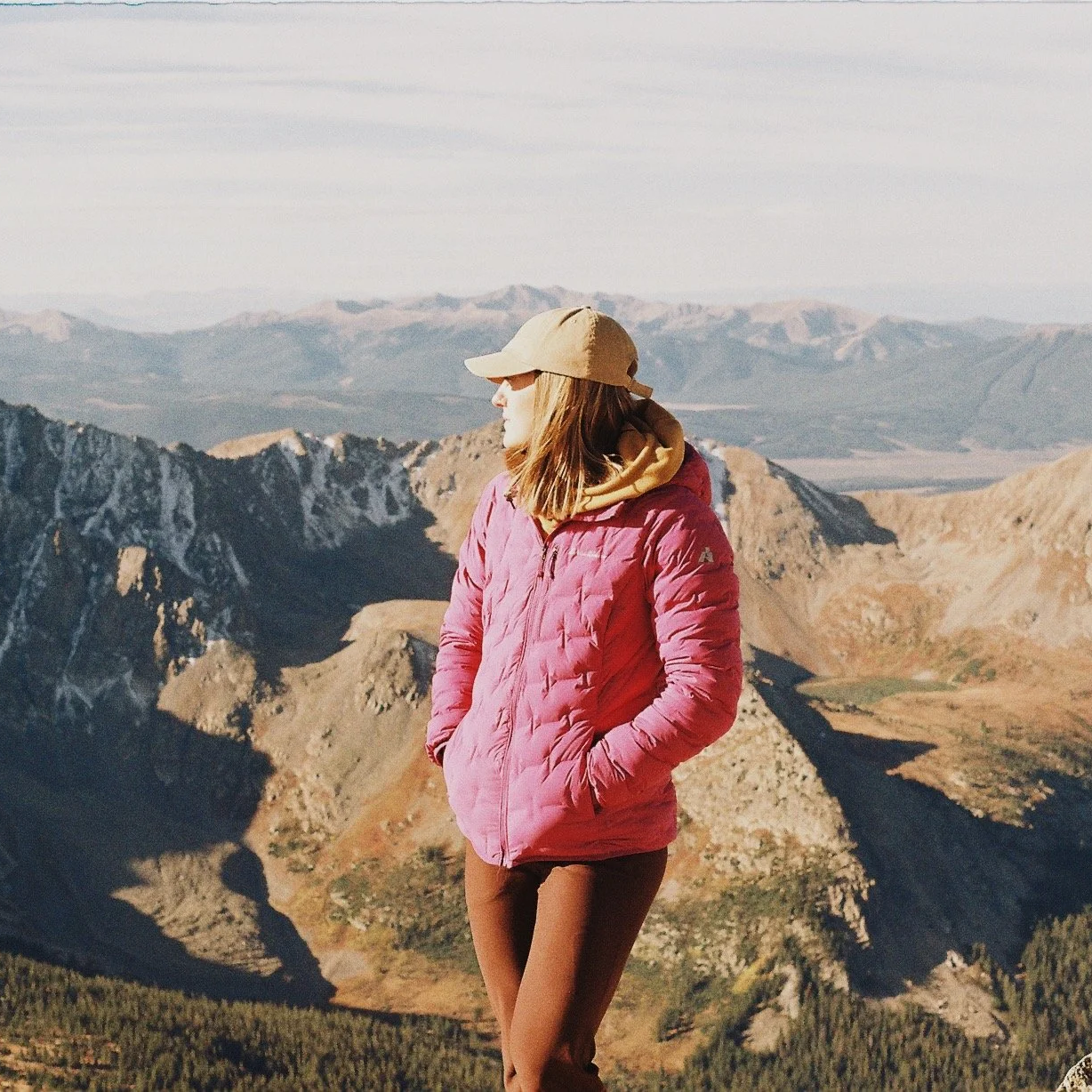 A woman in a pink jacket and a tan cap standing with her hands in her pockets on a mountain overlook with rugged peaks and valleys in the background.