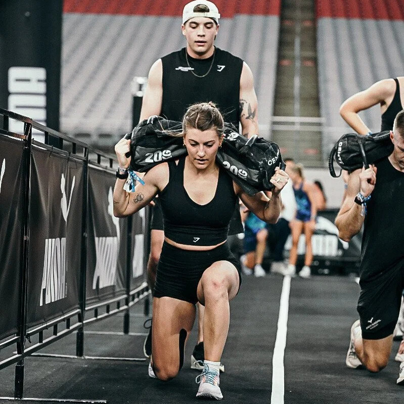 A woman doing a weighted lunge exercise at a gym with two men assisting her, one holding a sandbag on her shoulders and others in workout attire in the background.