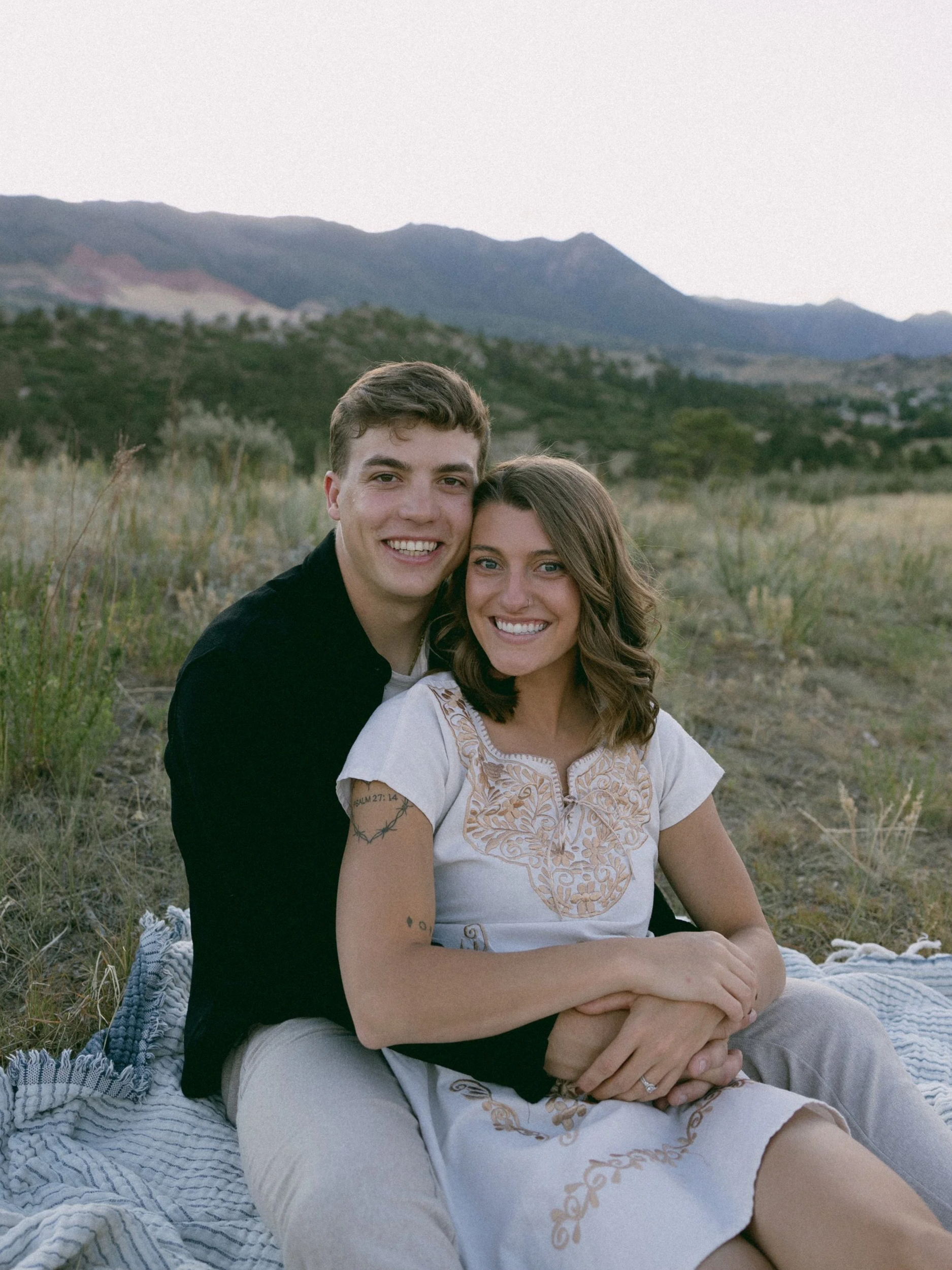 A young couple sitting on a blanket outdoors, smiling at the camera with mountains and green hills in the background.