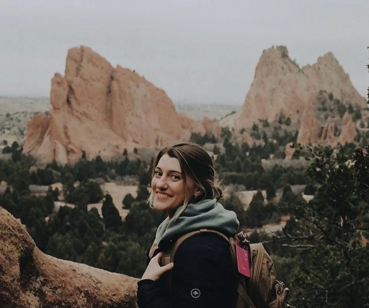 A woman smiling at the camera with a trail and desert landscape featuring large red rock formations in the background.