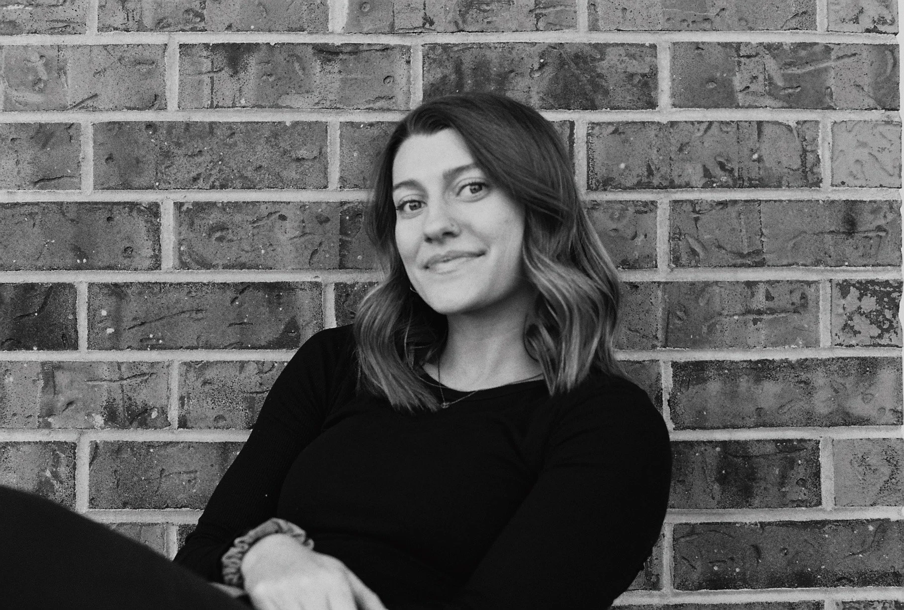 A woman with shoulder-length wavy hair sitting against a brick wall, smiling slightly, wearing a black top and a watch on her left wrist.