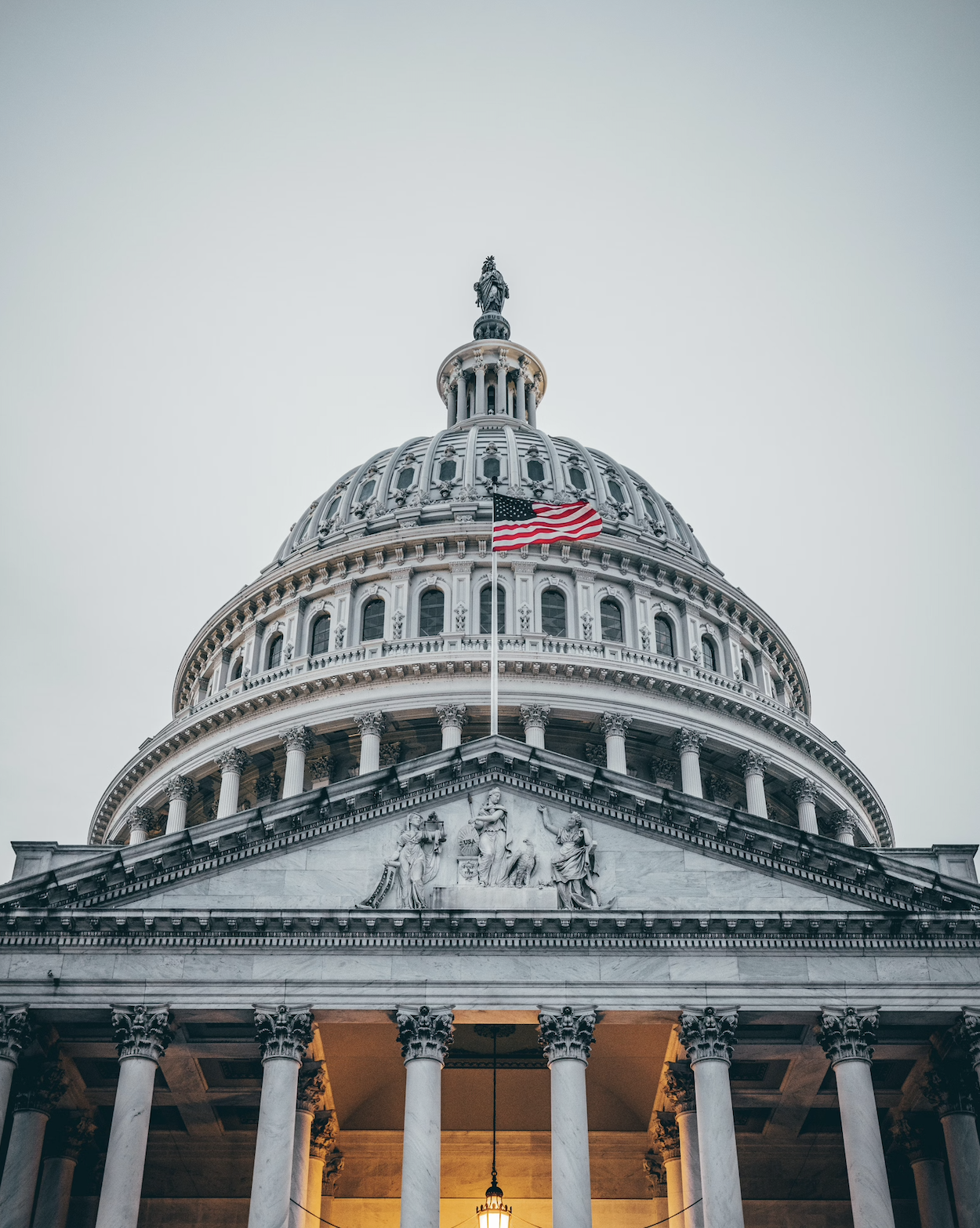 Close-up of the United States Capitol building dome with an American flag, classical architectural details, and statues, illuminated from below.