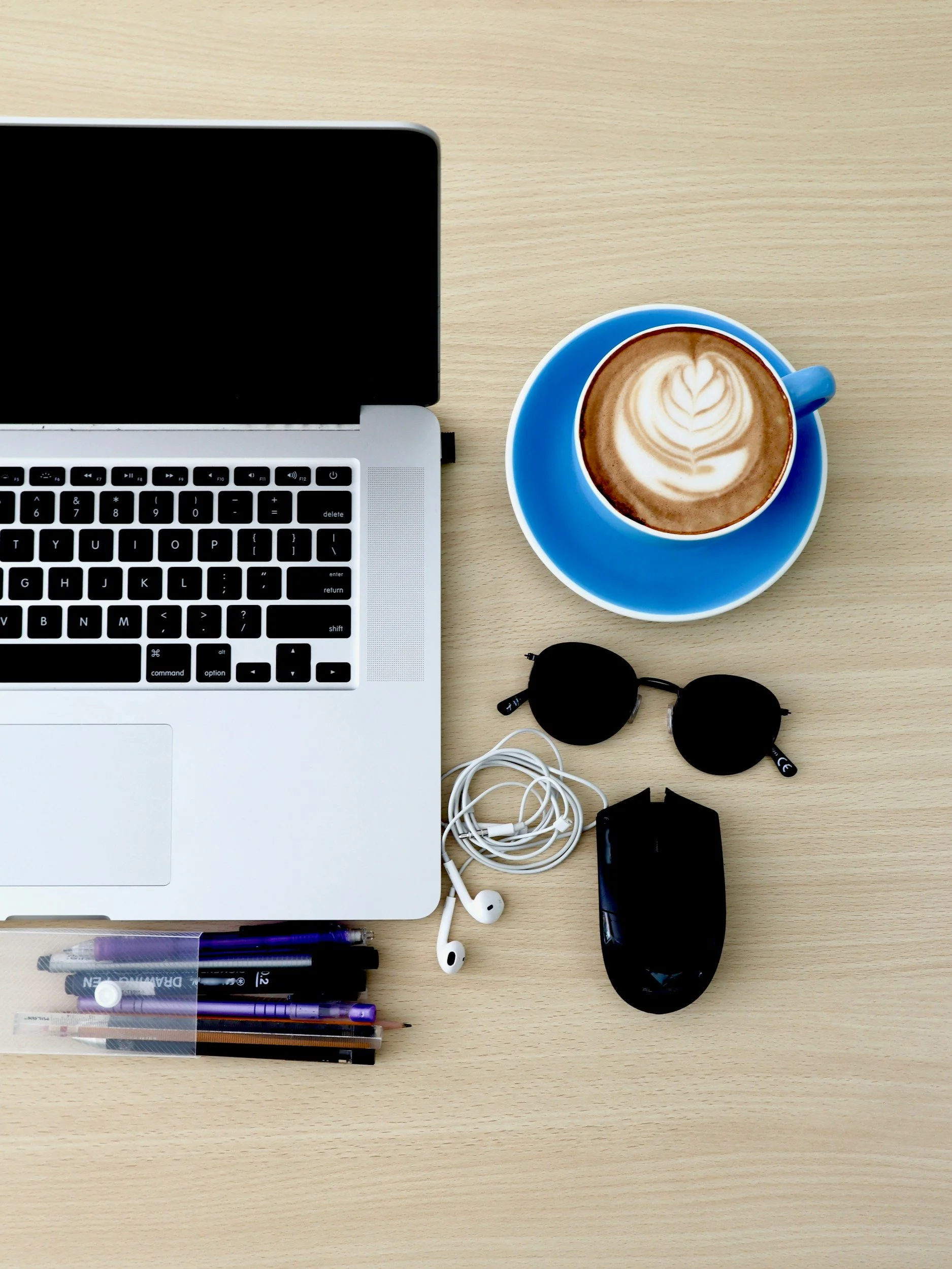 Laptop, cup of coffee with latte art on a wooden desk, sunglasses, earphones, black electronic device, set of pens and markers, and some markers in a clear case.