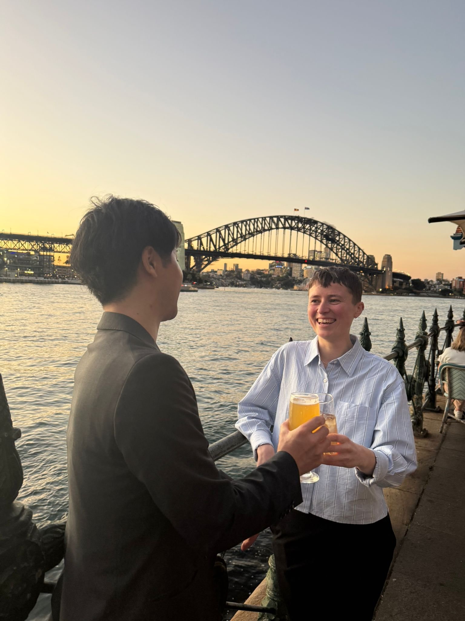 A man and his English tutor are standing near a railing by a river, smiling and holding glasses of beer, with the Sydney Harbour Bridge in the background at sunset.