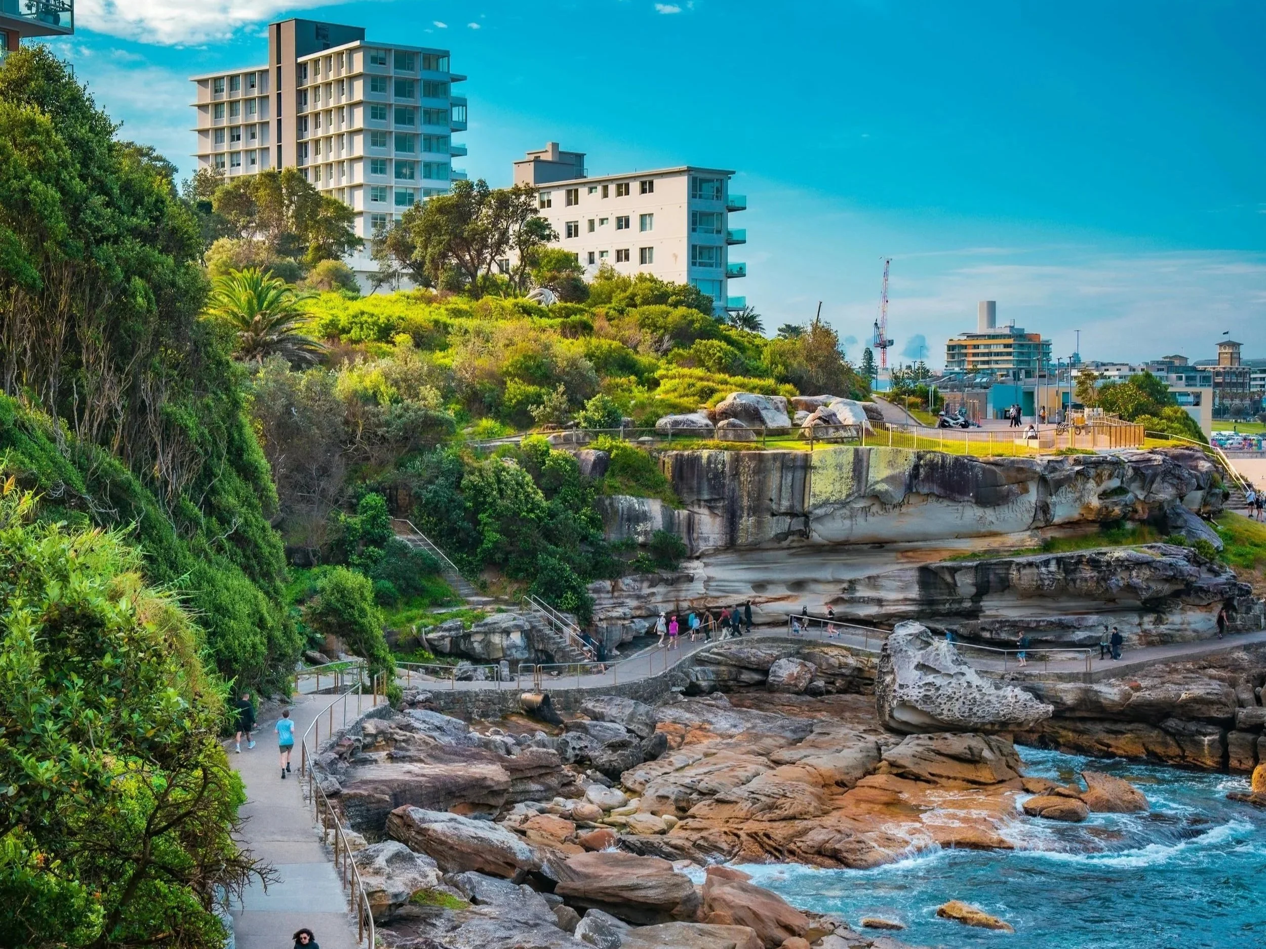 A coastal walkway with people walking, overlooking rocky shoreline and ocean, with modern buildings and green trees in the background on a clear day.