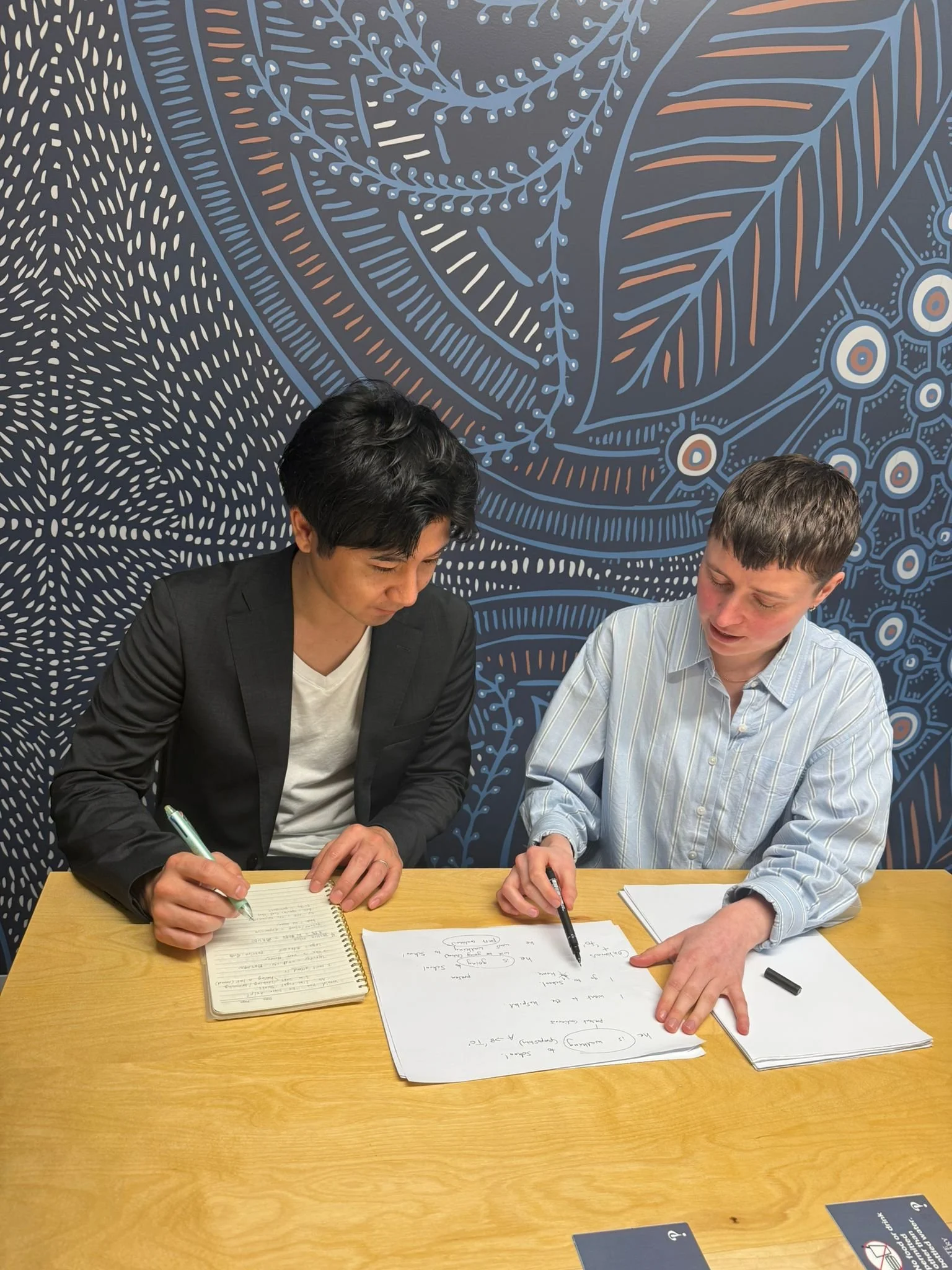 A man and a woman are sitting at a wooden table, learning English and taking notes. They are engaged in a discussion, with a blue patterned wall in the background.
