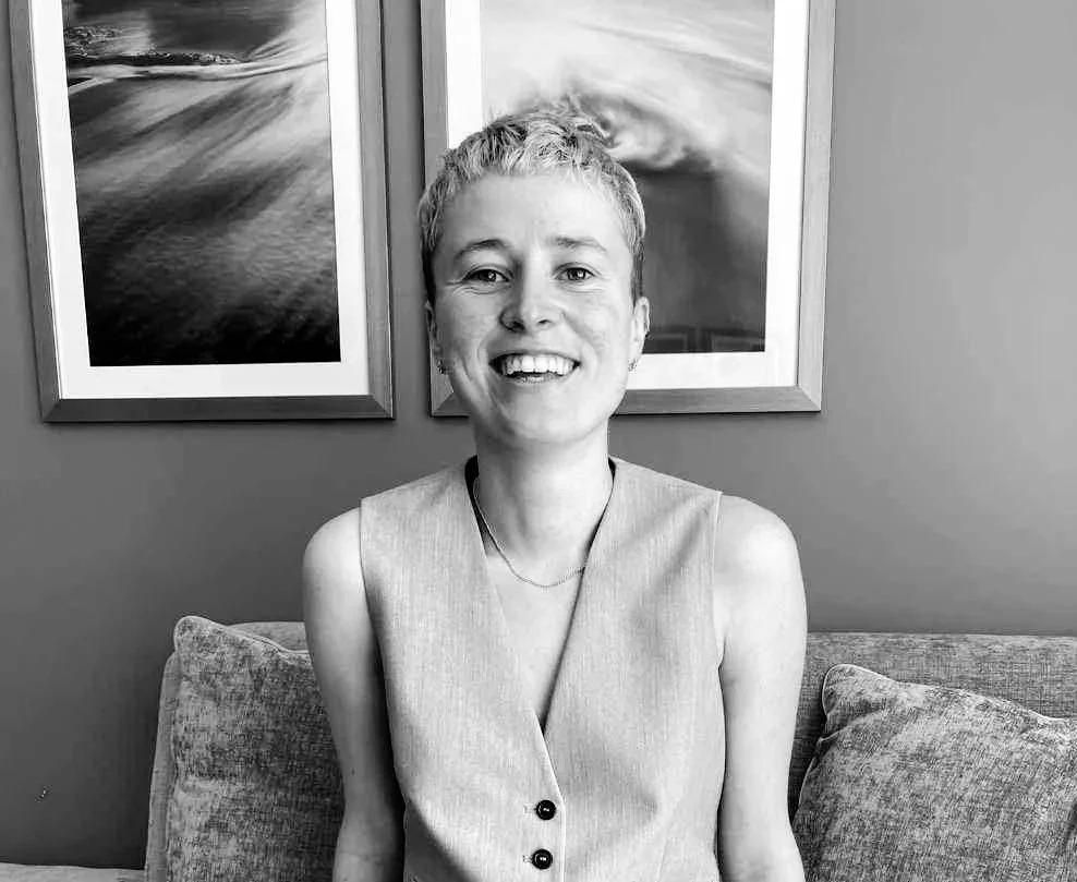 A smiling woman with short hair sitting on a couch in front of two framed landscape photographs on a wall.