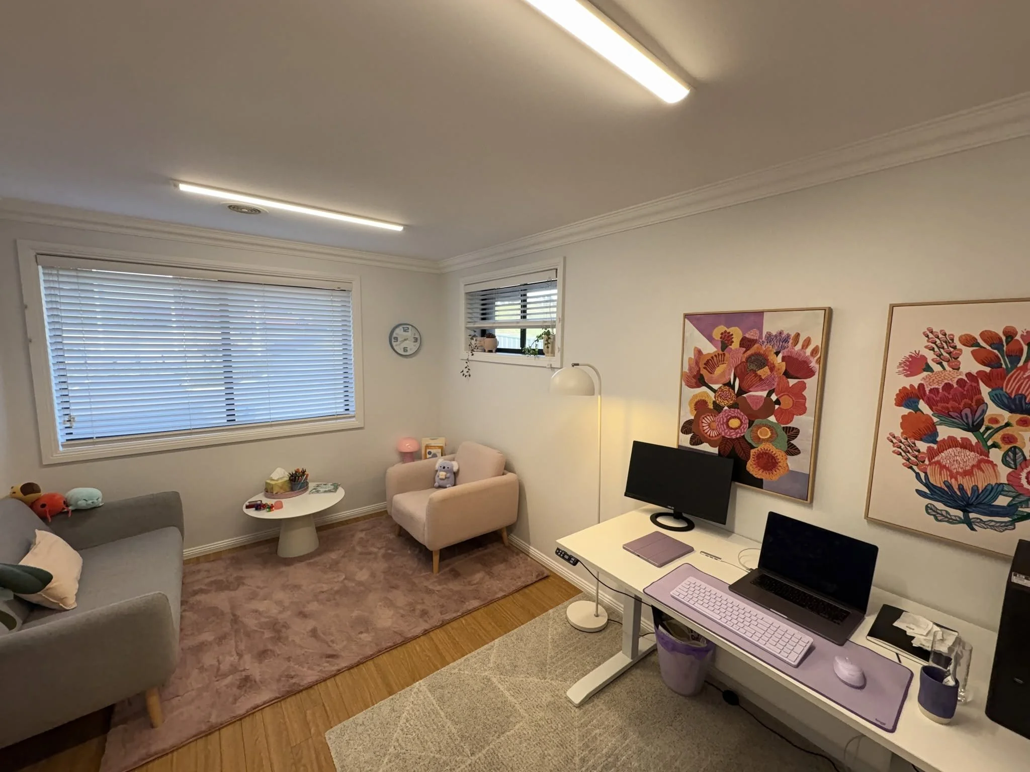 A cozy living room with a beige armchair, a small white side table, a gray sofa with plush toys, a computer desk with a laptop, monitor, and pink accessories, colorful floral artwork on the wall, and large windows with white blinds, illuminated by fluorescent ceiling lights.