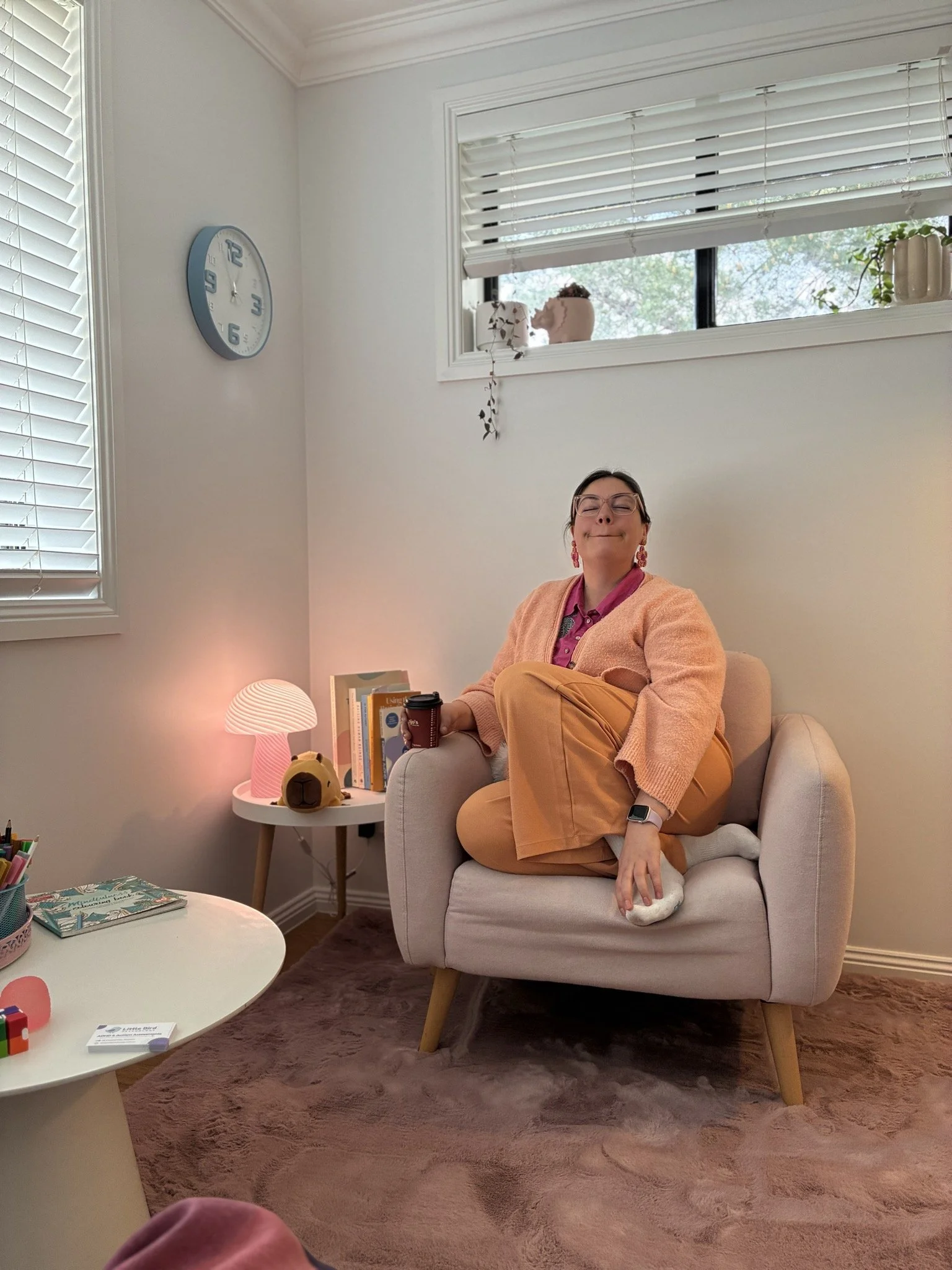 A woman sitting cross-legged on a beige armchair in a living room. She is smiling with her eyes closed, wearing glasses, a pink shirt, an orange sweater, and tan pants. There is a white side table with books, a black and red travel mug, and a yellow plush toy. A pink lamp and some art supplies are on a table in the foreground. The room has white walls, a large window with white blinds, and a pink carpet.