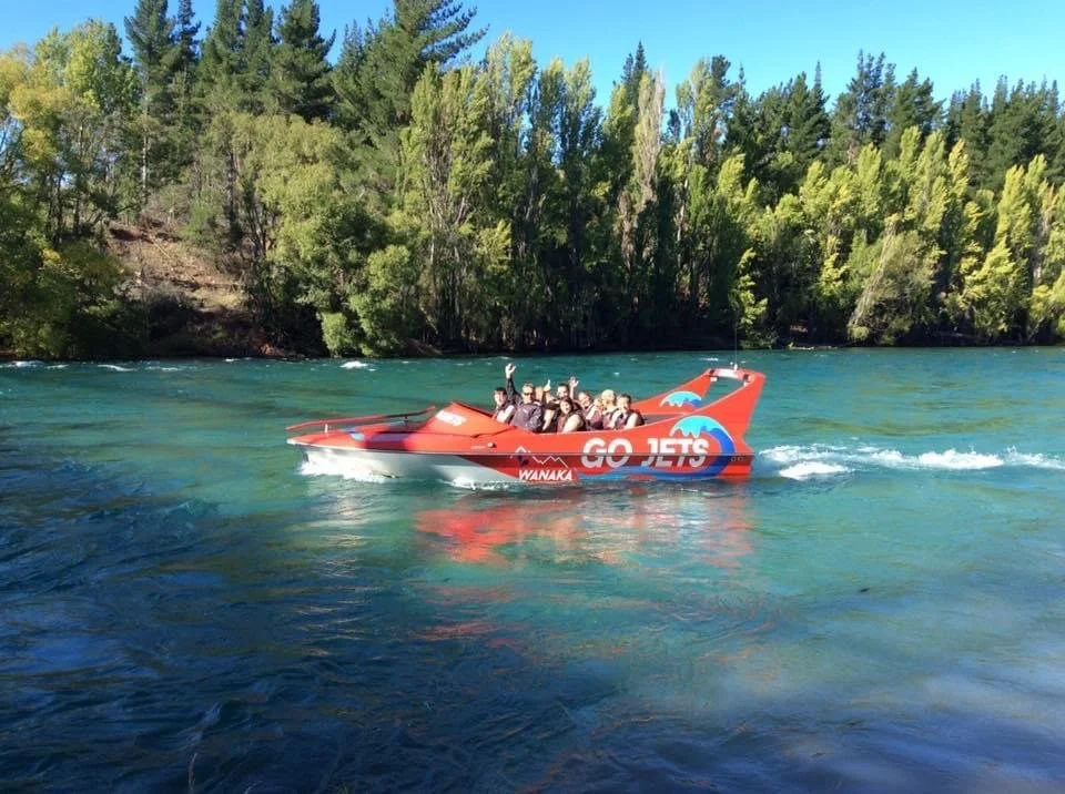 A group of people on a red and white jet boat on a river with green trees in the background.
