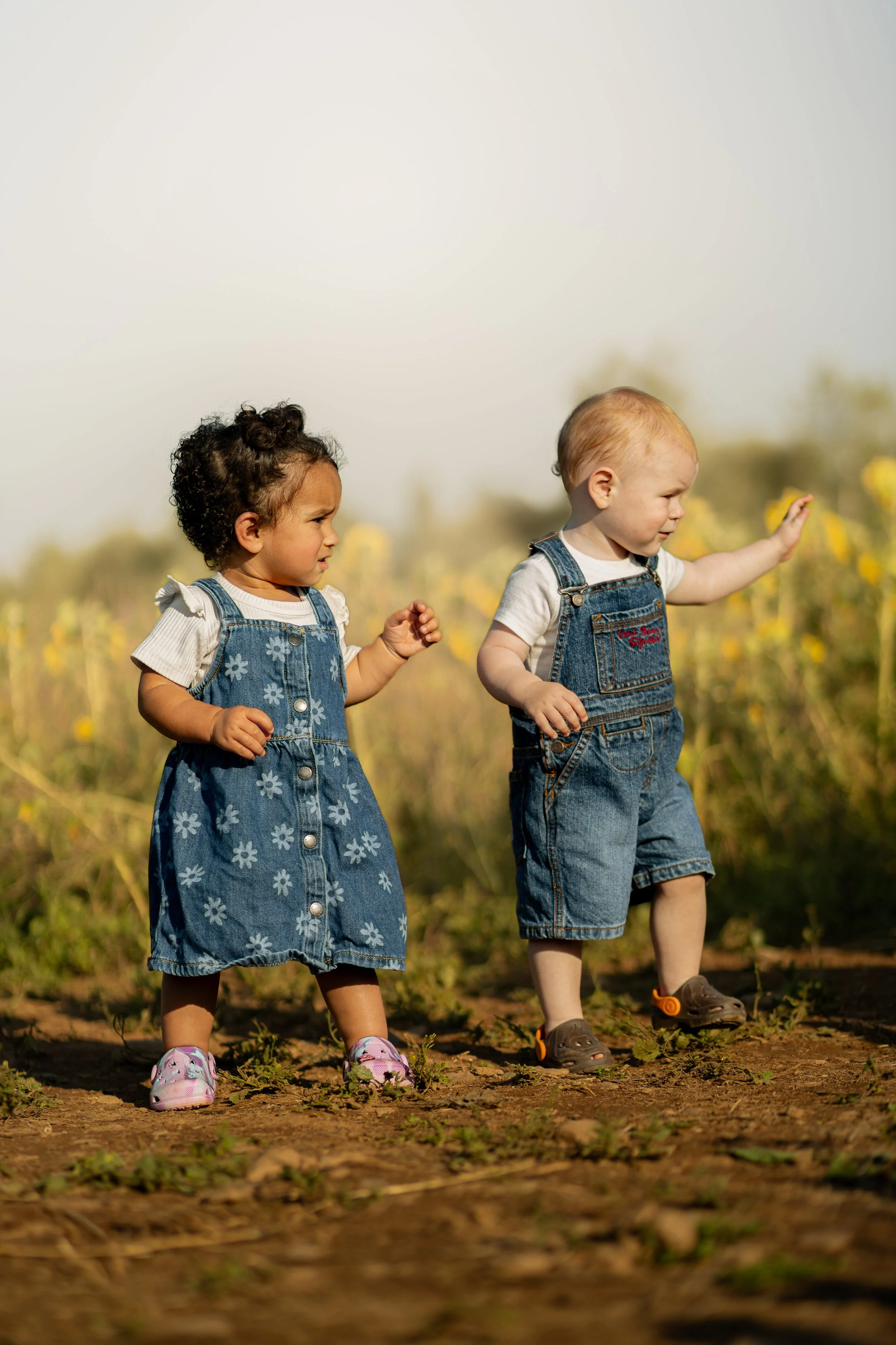 Two young children, a girl with curly hair and a boy with straight hair, walk outdoors on a dirt path surrounded by yellow flowers, wearing denim overalls and casual shoes.