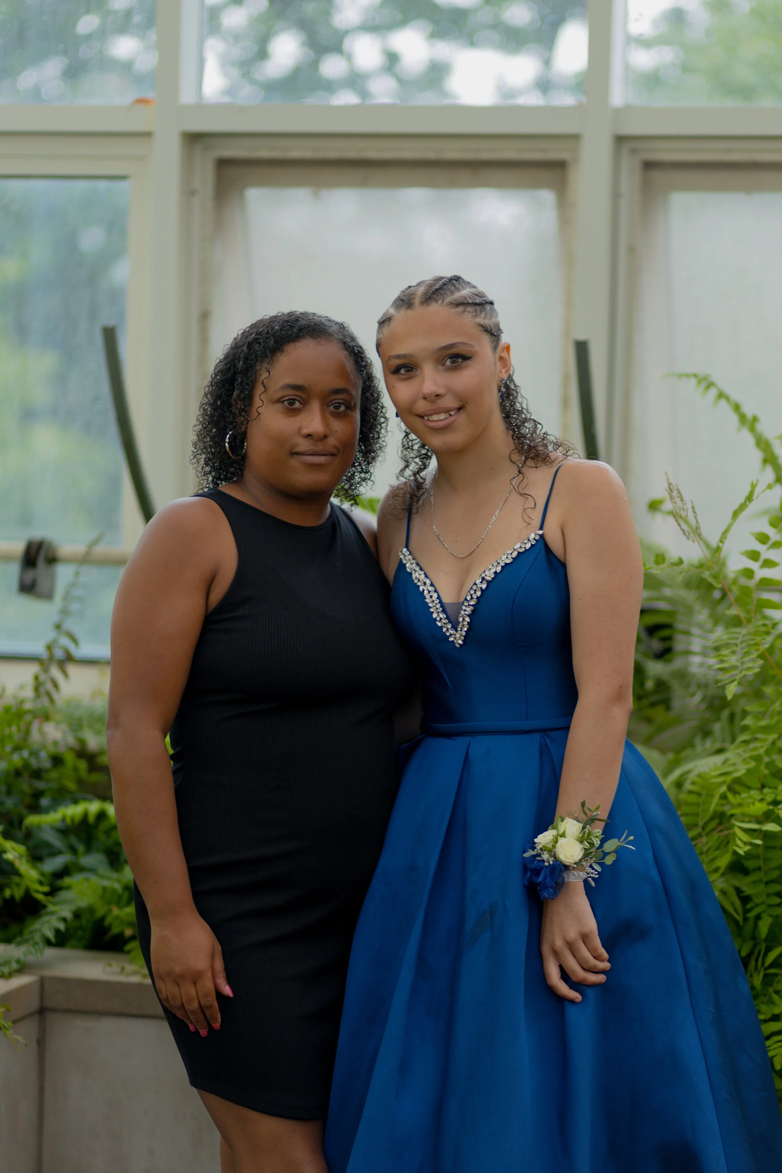 Two women standing close together in front of large windows with greenery outside. One woman wears a black sleeveless dress, and the other wears a blue gown with a corsage on her wrist.