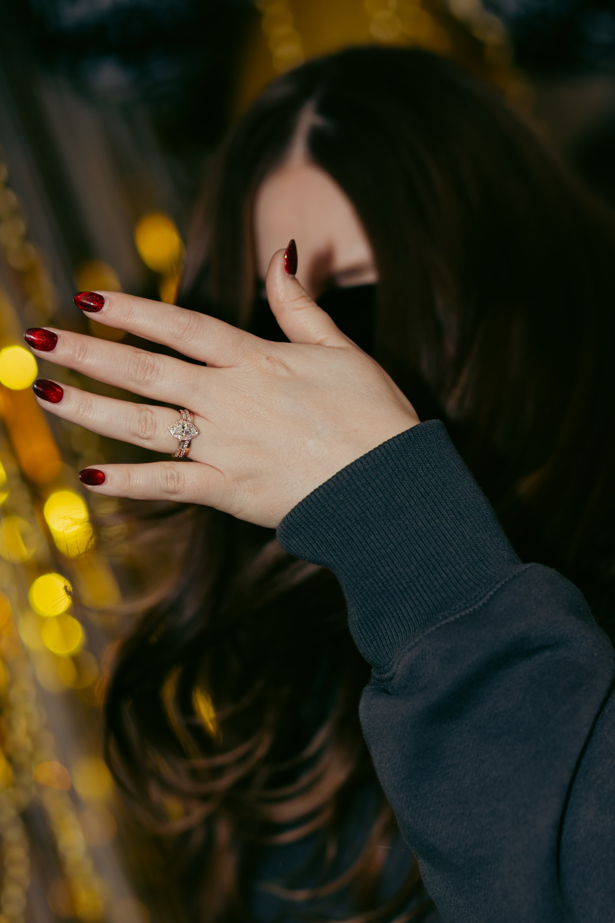 A person showing off a diamond engagement ring on their finger, with red nail polish, covering part of their face with their hand, wearing a dark long-sleeve shirt, with warm yellow out-of-focus lights in the background.