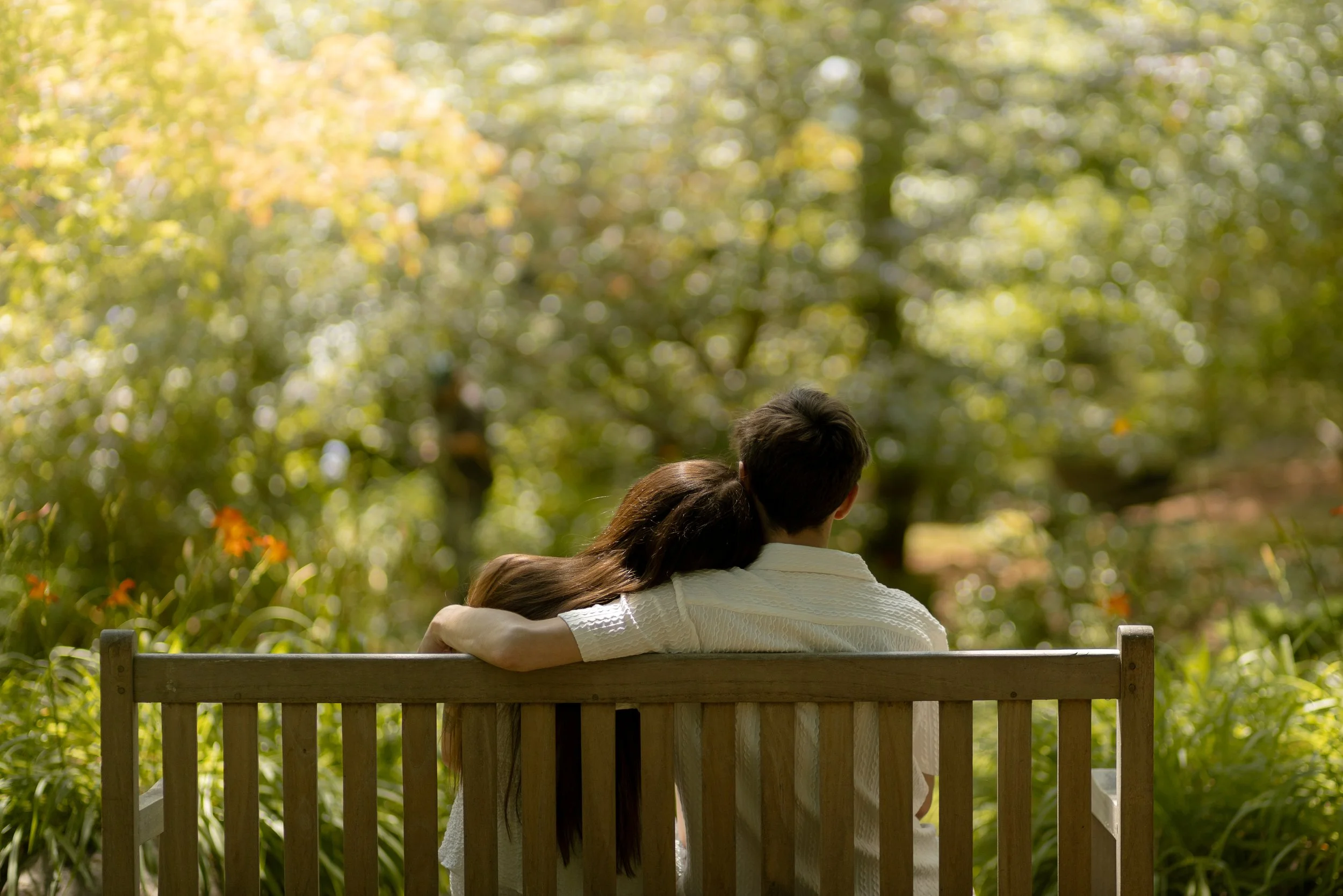 Two people sitting on a wooden bench in a park with trees and foliage in the background, embracing while looking away from the camera.