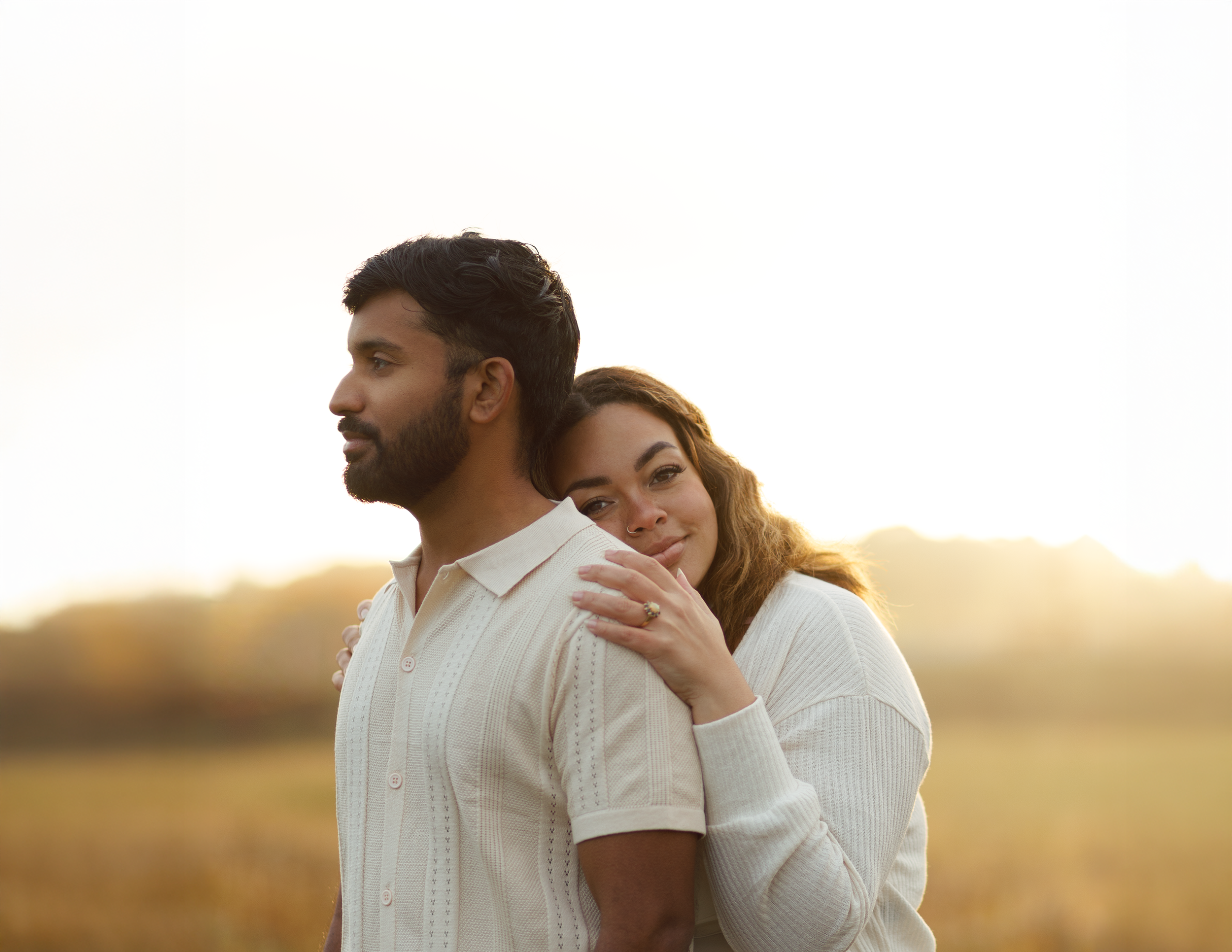 A woman with curly hair rests her head on a man's shoulder, both standing outdoors during sunset. The man has dark hair and a beard, and is looking to the side. The woman is smiling softly with her eyes slightly closed, embracing the man with her han