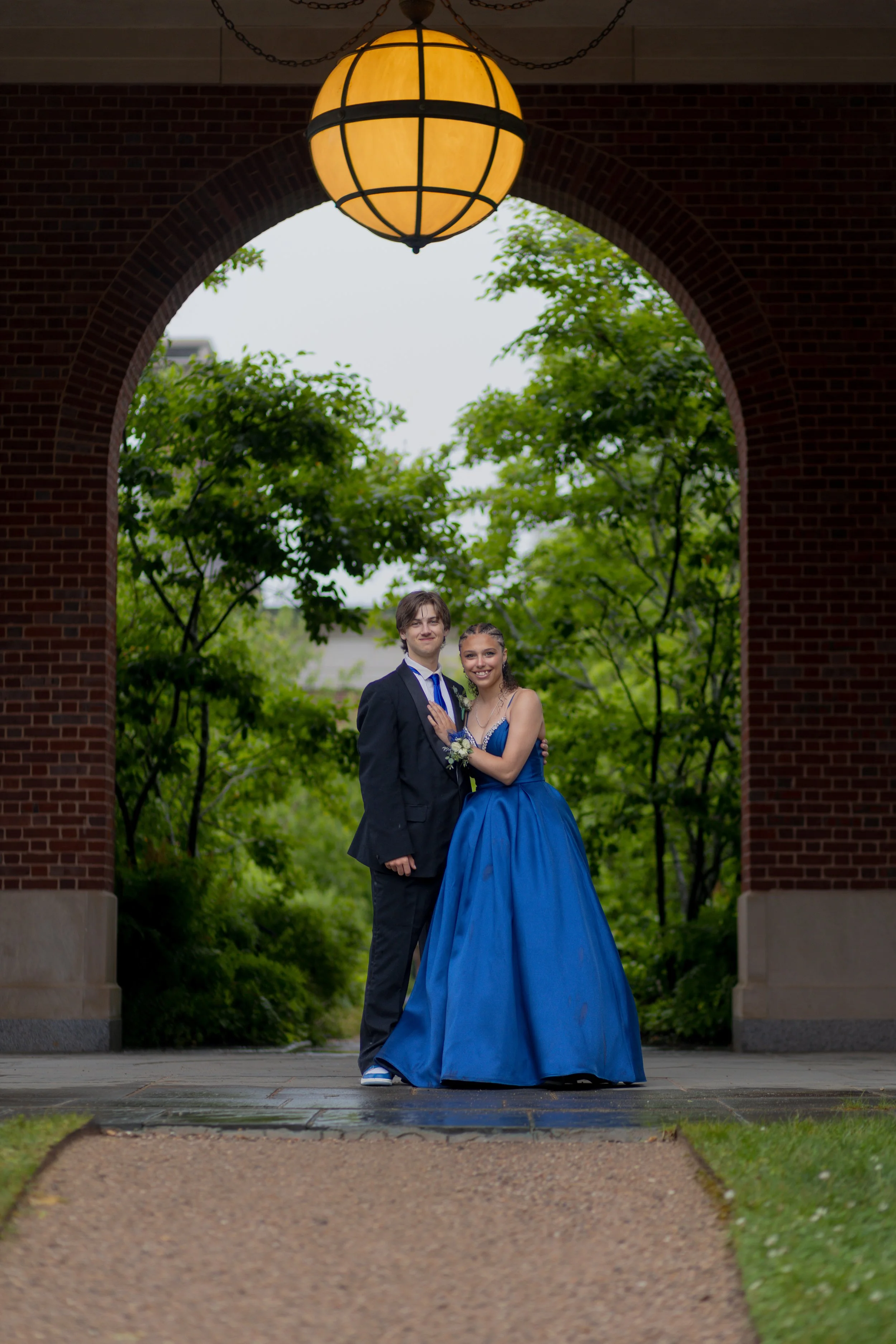 A young couple dressed formally, standing under a brick archway, with greenery in the background and a large hanging yellow lamp overhead.