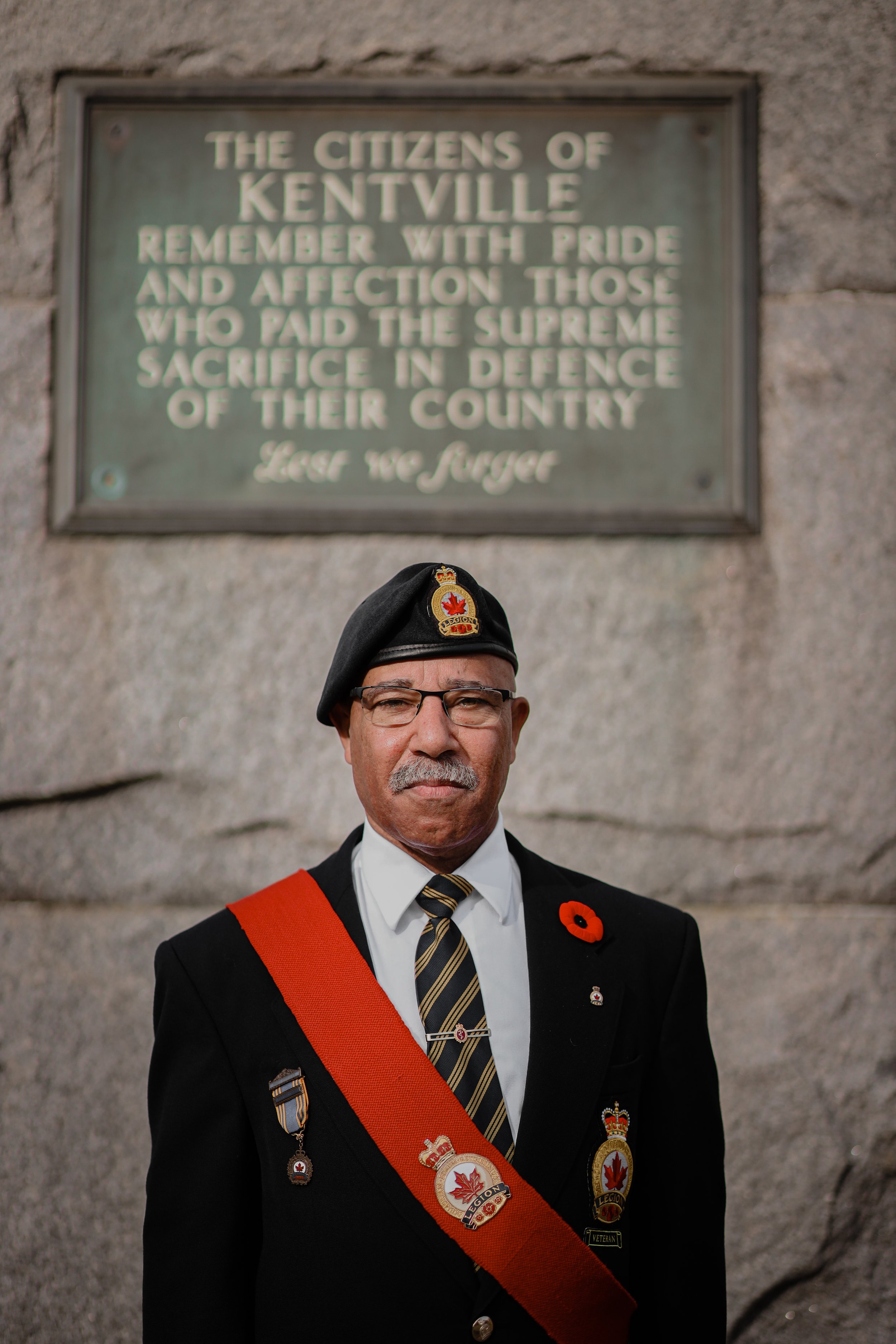 A man dressed in military uniform standing in front of a memorial plaque with the text "The citizens of Kentville remember with pride and affection those who paid the supreme sacrifice in defence of their country."