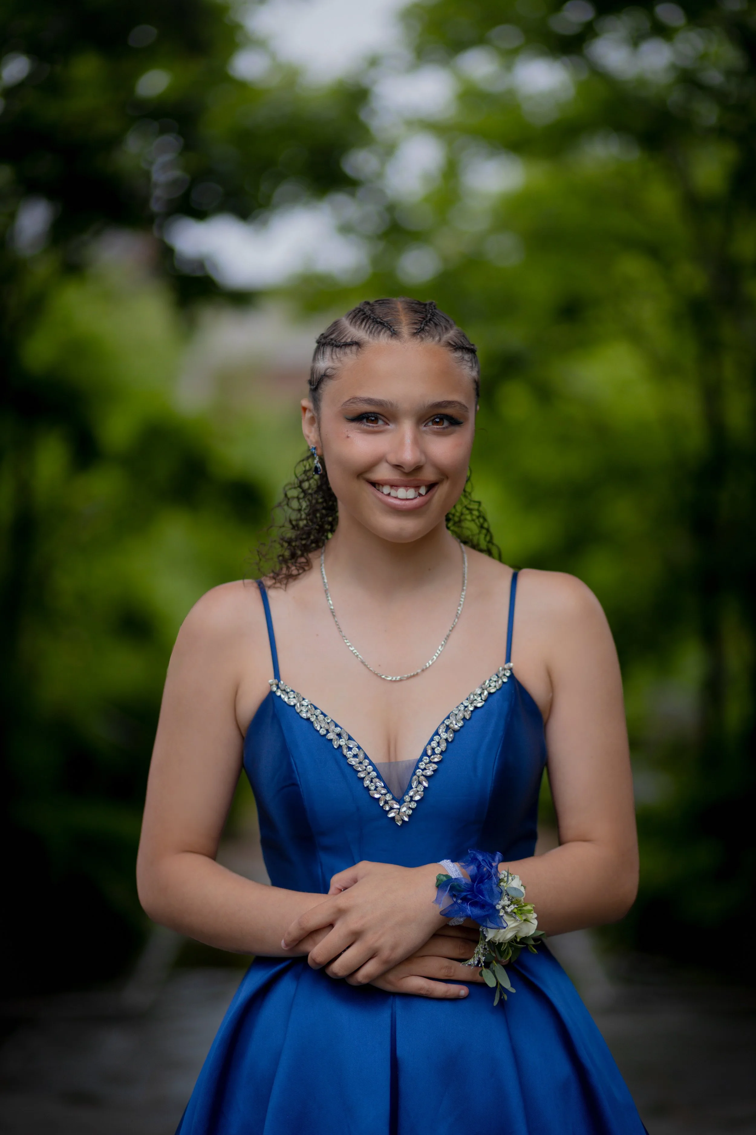 A young woman with curly hair styled in braids, wearing a royal blue formal dress with silver embellishments, standing outdoors with a blurred green background, smiling at the camera, with a corsage on her wrist.