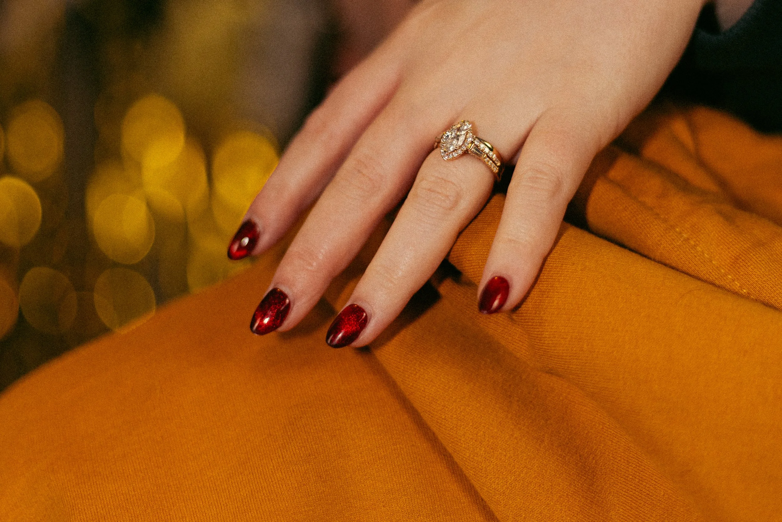 A close-up of a woman's hand with dark red, glittery nail polish, wearing a gold ring with a large, oval-shaped diamond surrounded by smaller diamonds on her ring finger. The hand rests on mustard-colored fabric, with a blurred background of golden l