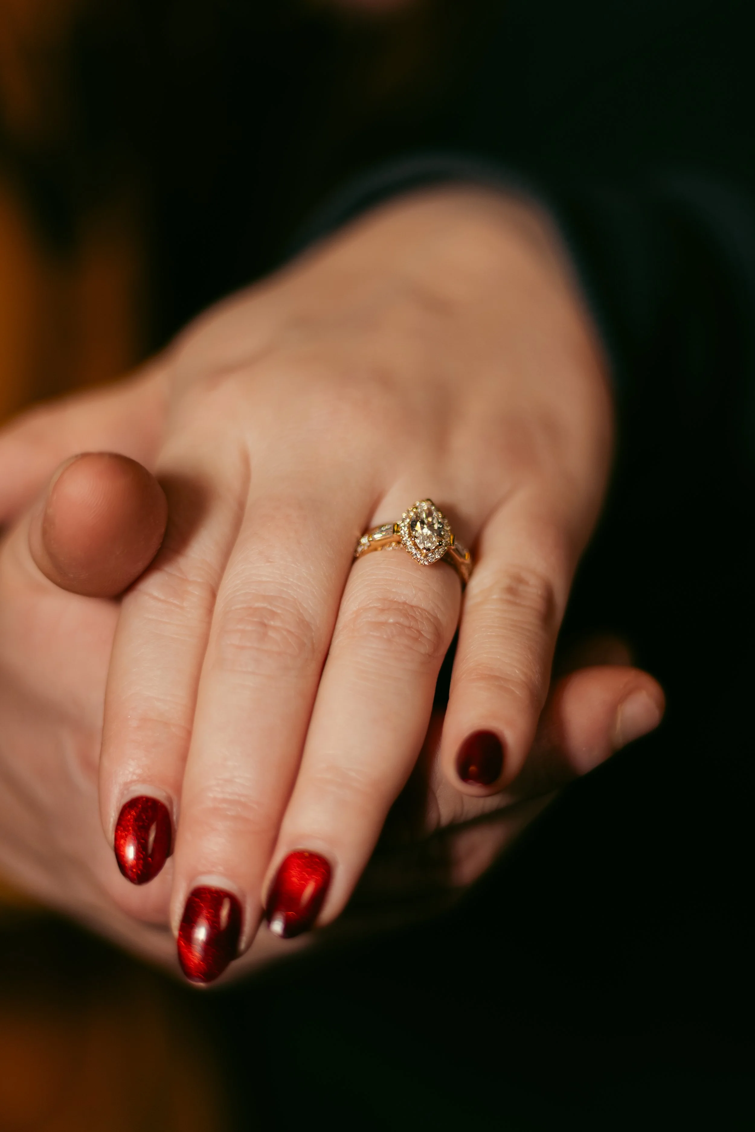 A person's hand with red metallic nail polish, wearing a gold ring with a large oval-shaped diamond surrounded by smaller diamonds.