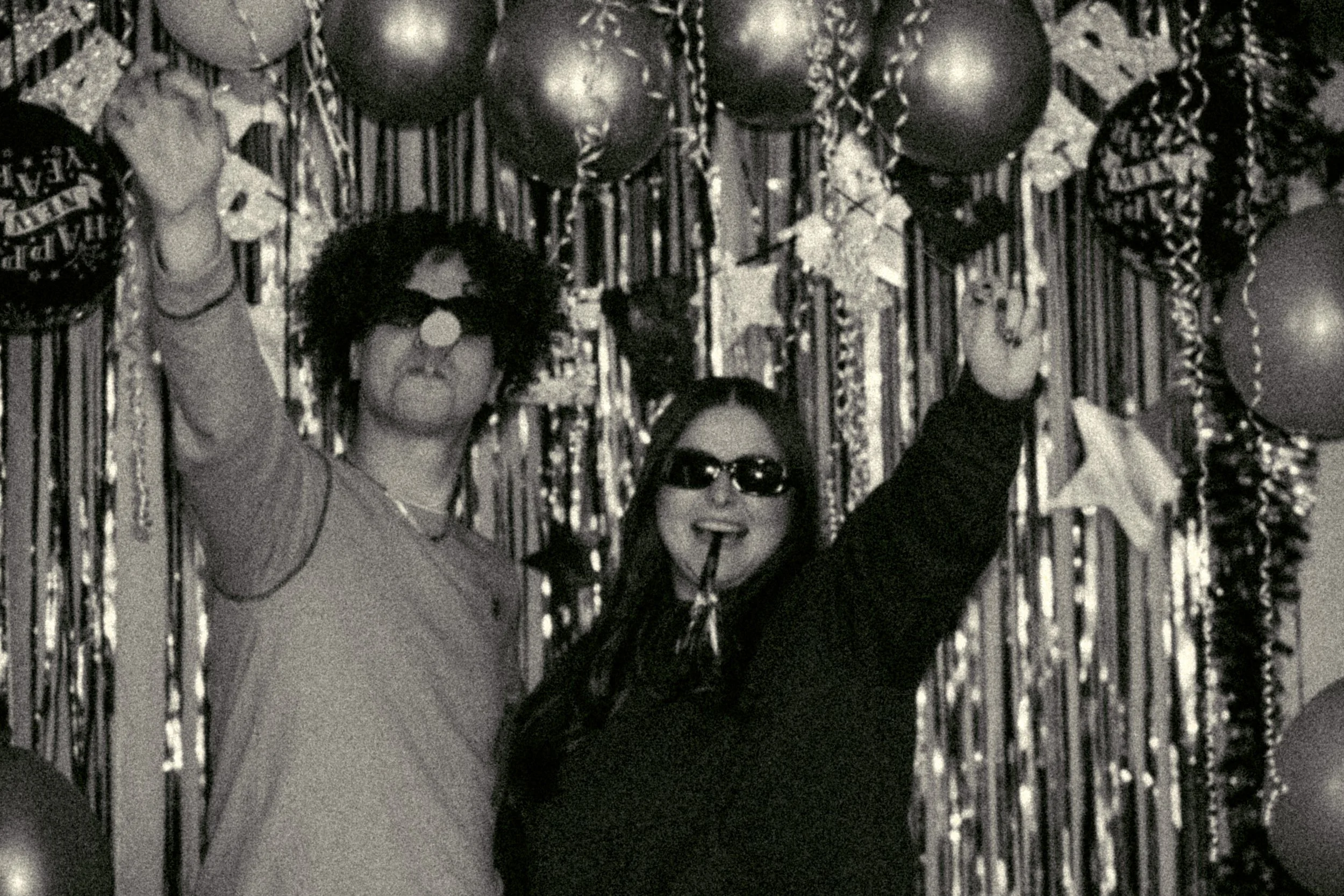 Two women celebrating at a party, wearing sunglasses and party hats, with balloons and streamers in the background.