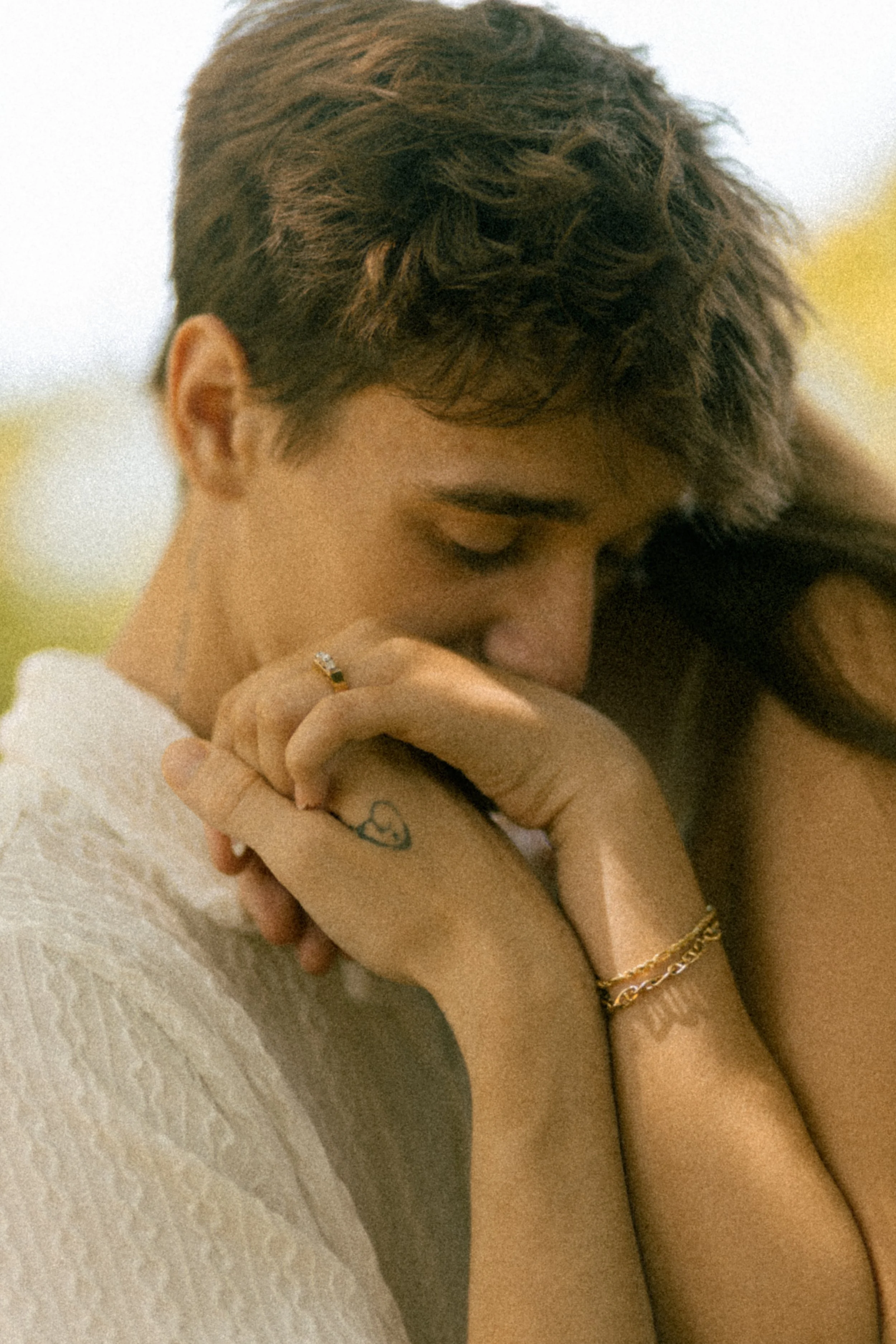 A person with short hair holds another person's hand close to their face, eyes closed, in a tender moment. The person has a ring, a small tattoo, and gold bracelets.