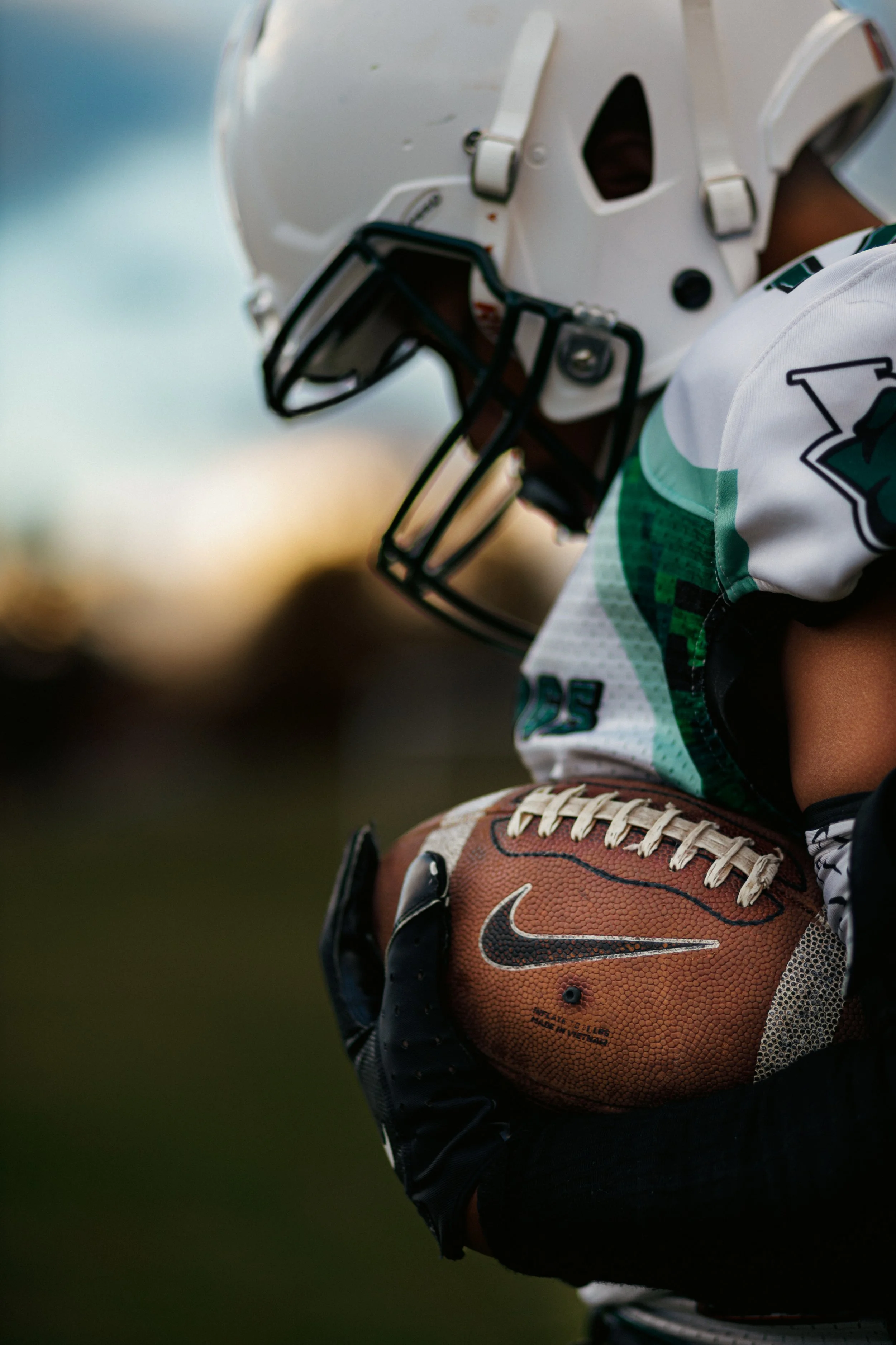 Close-up of an American football player holding a football, wearing a white helmet and jersey with green accents, and black gloves.