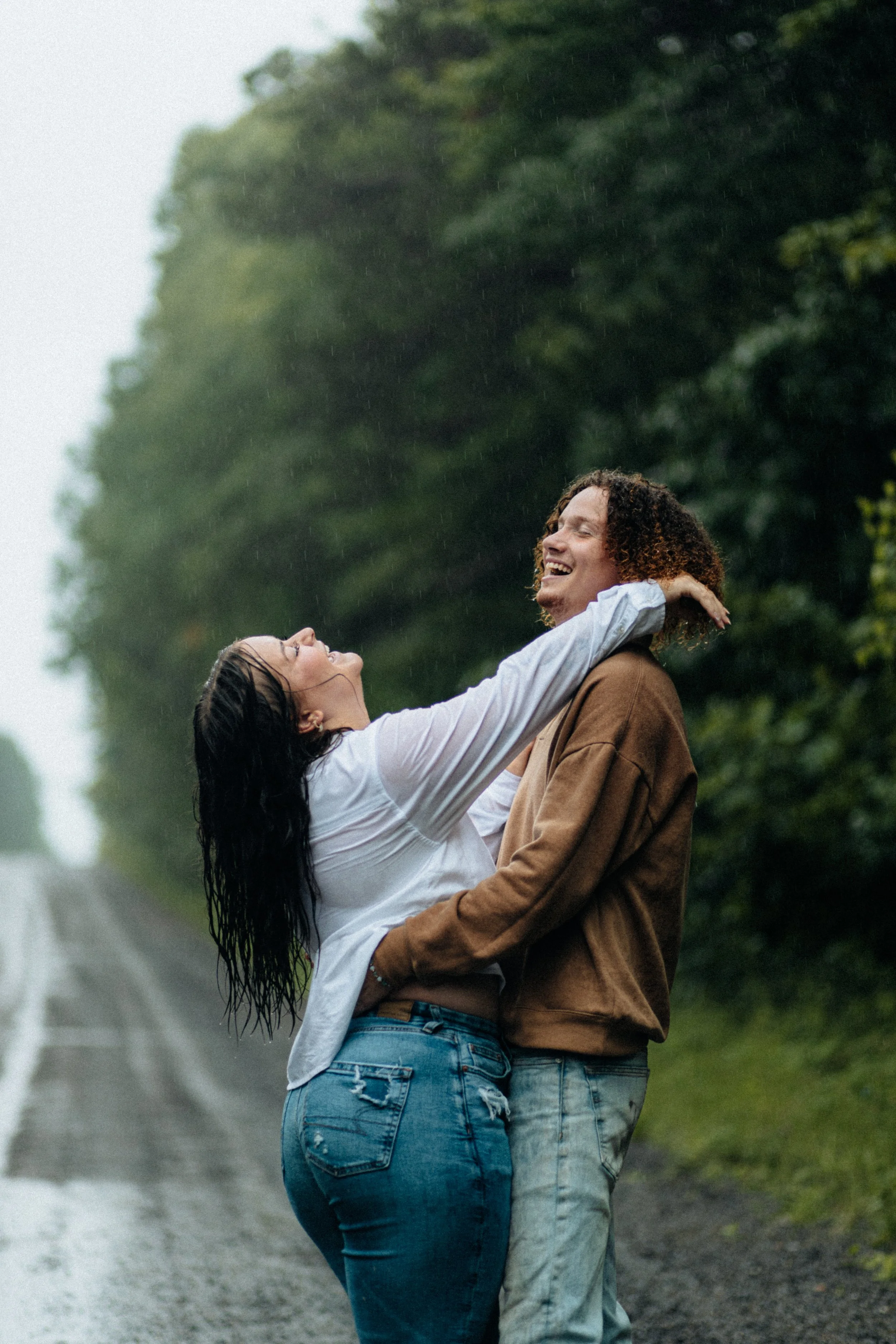 A couple happily embracing in the rain on a forested roadside, smiling and enjoying each other's company.