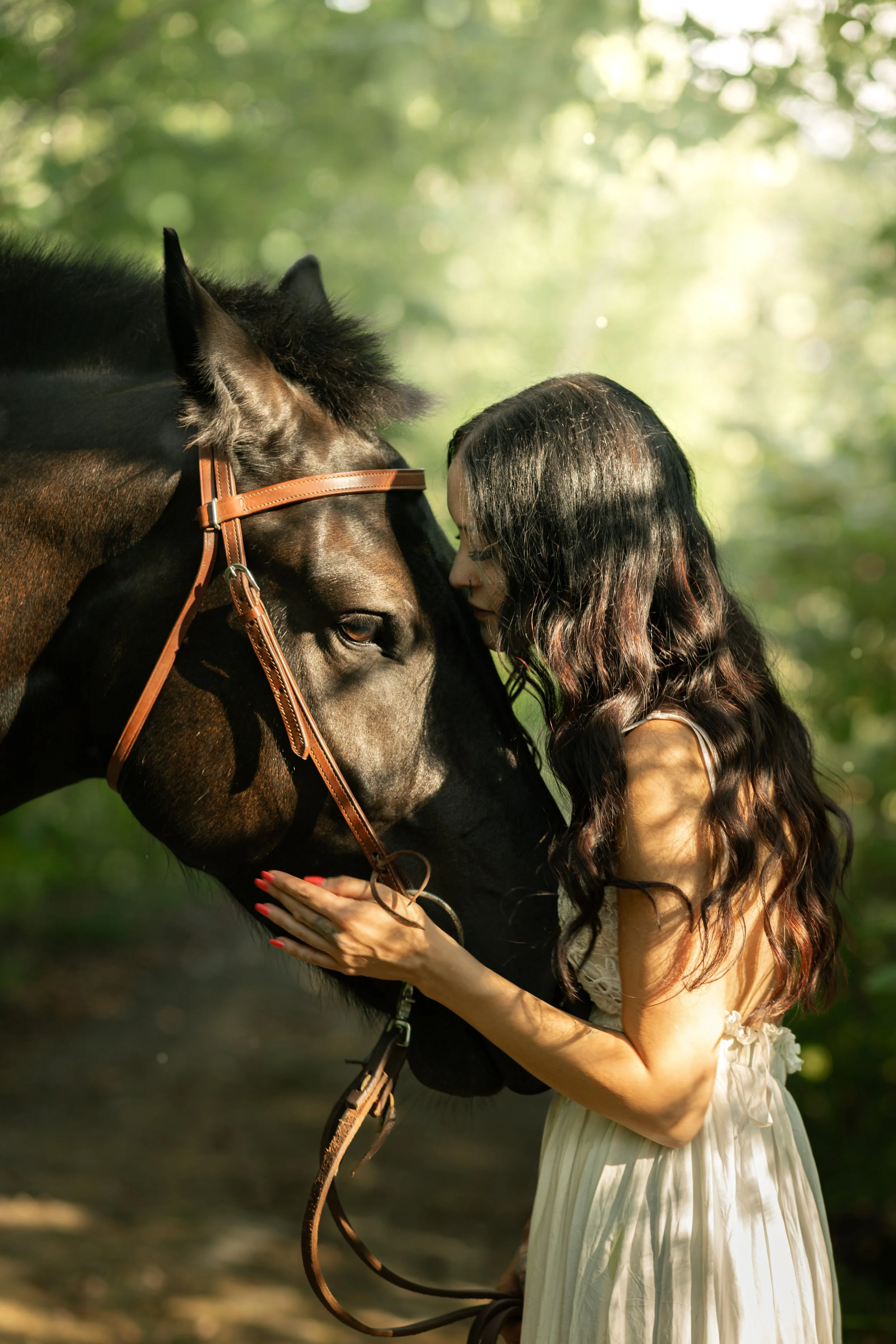 A woman with long wavy dark hair and a beige dress gently touches and nuzzles a dark brown horse with a leather bridle, in a lush green outdoor setting.