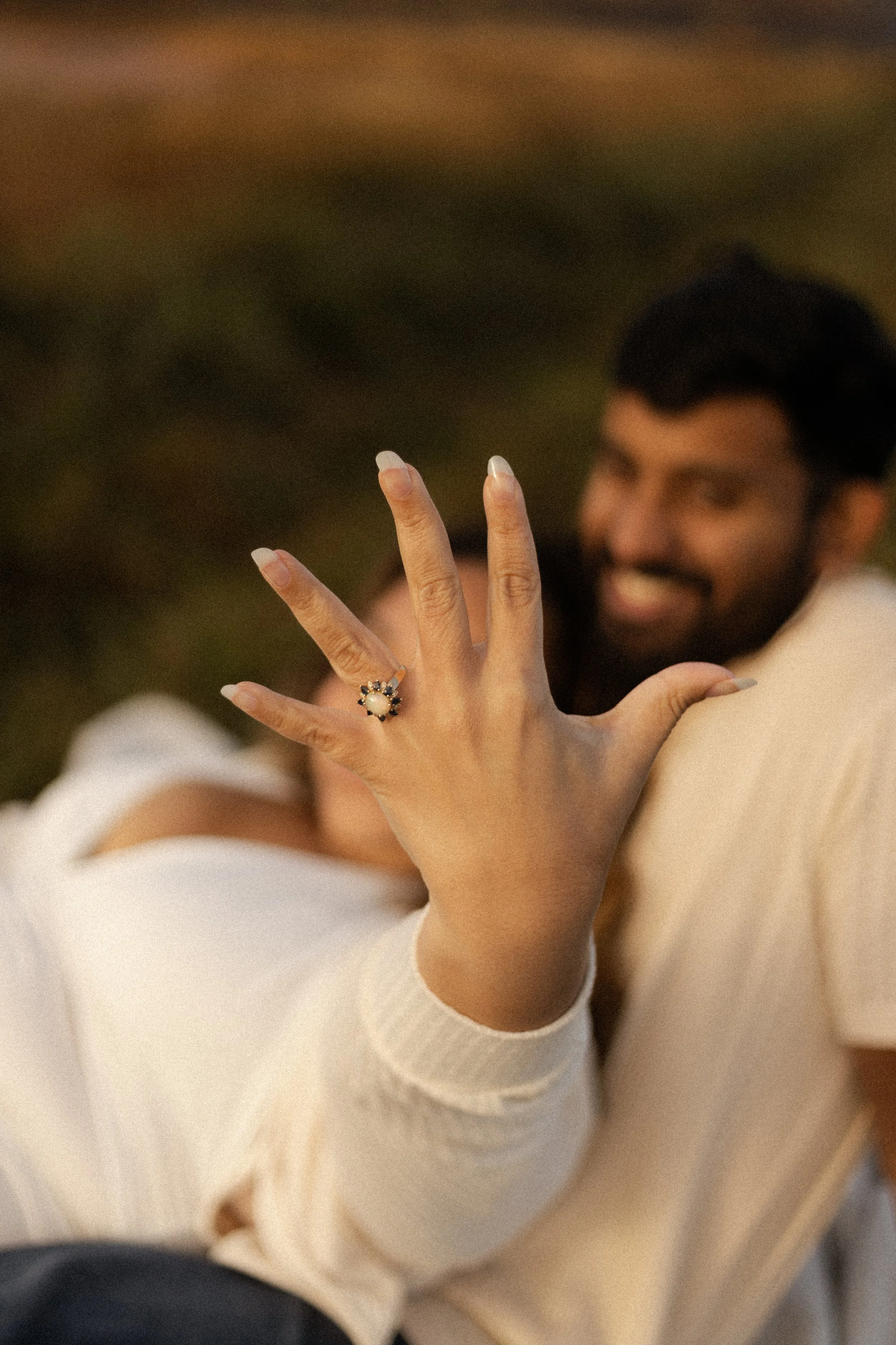 A woman showing off her engagement ring on her finger while sitting with a man who is smiling, both are outdoors during sunset.