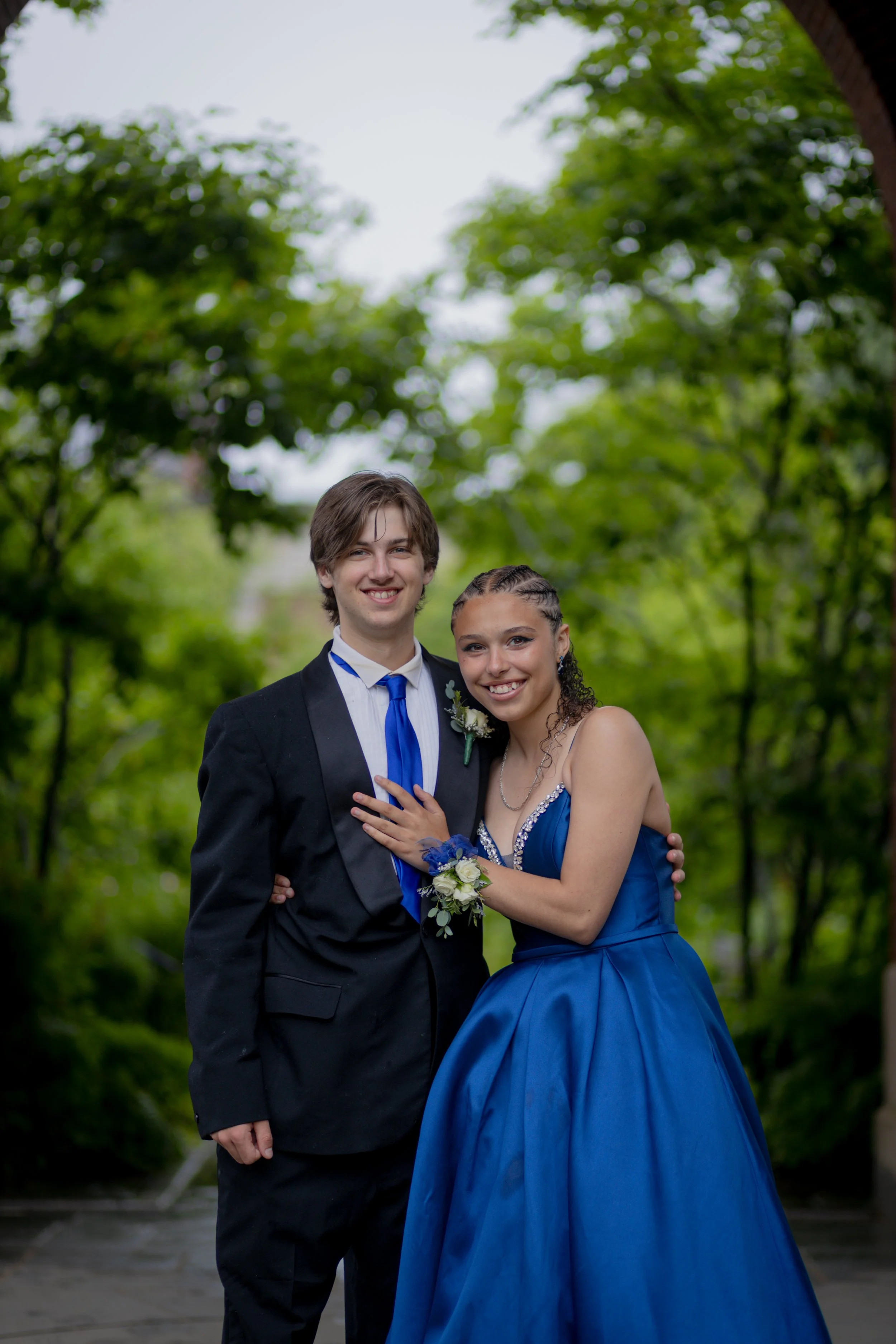 Young couple dressed in formal attire, smiling outdoors on a cloudy day, with greenery in the background.
