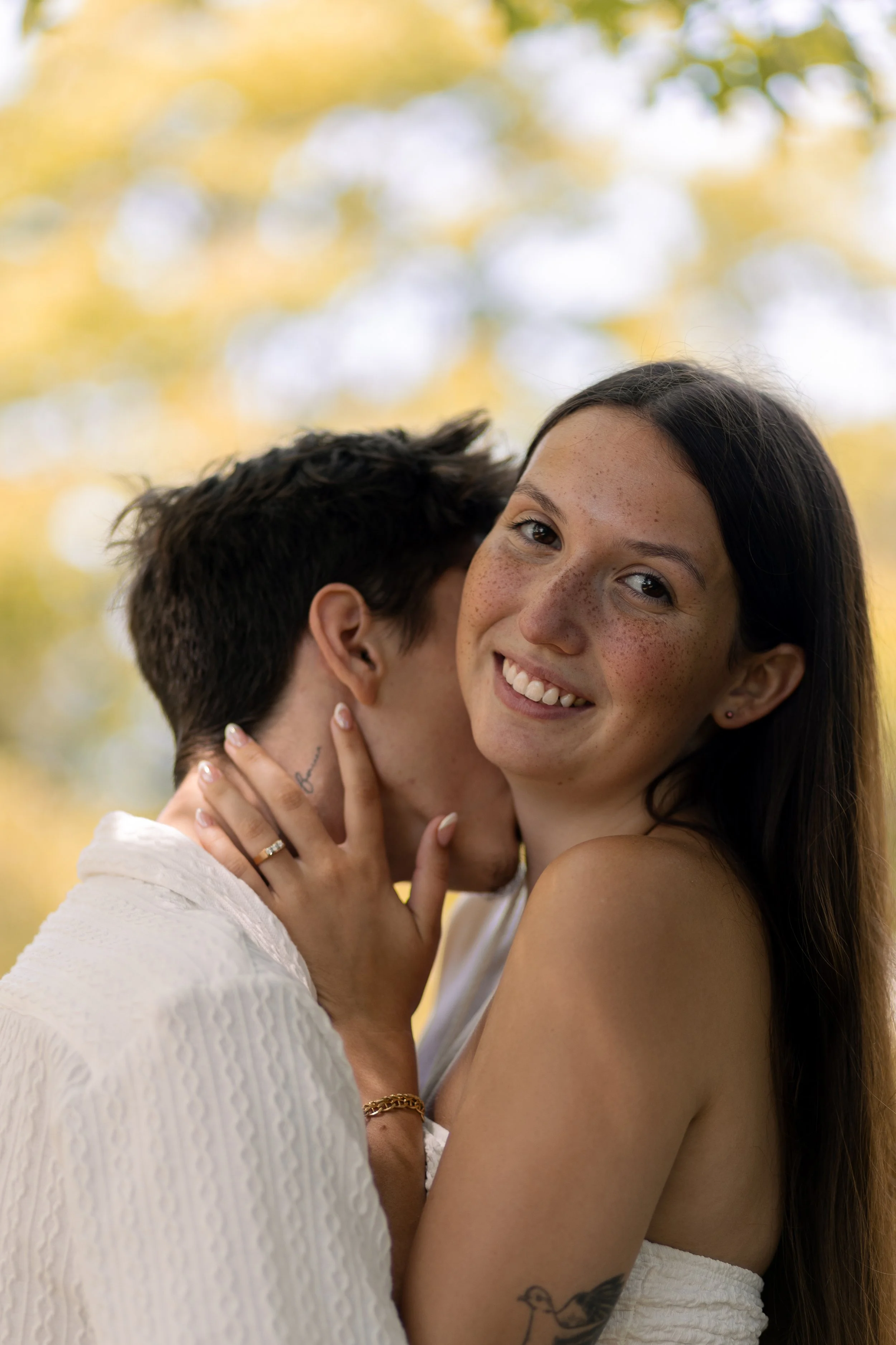 A smiling woman with freckles and dark hair embraces a man with short dark hair, who is whispering in her ear. The woman is wearing a white strapless top, a gold bracelet, and a ring. They are outdoors during daytime with a blurred background of tree